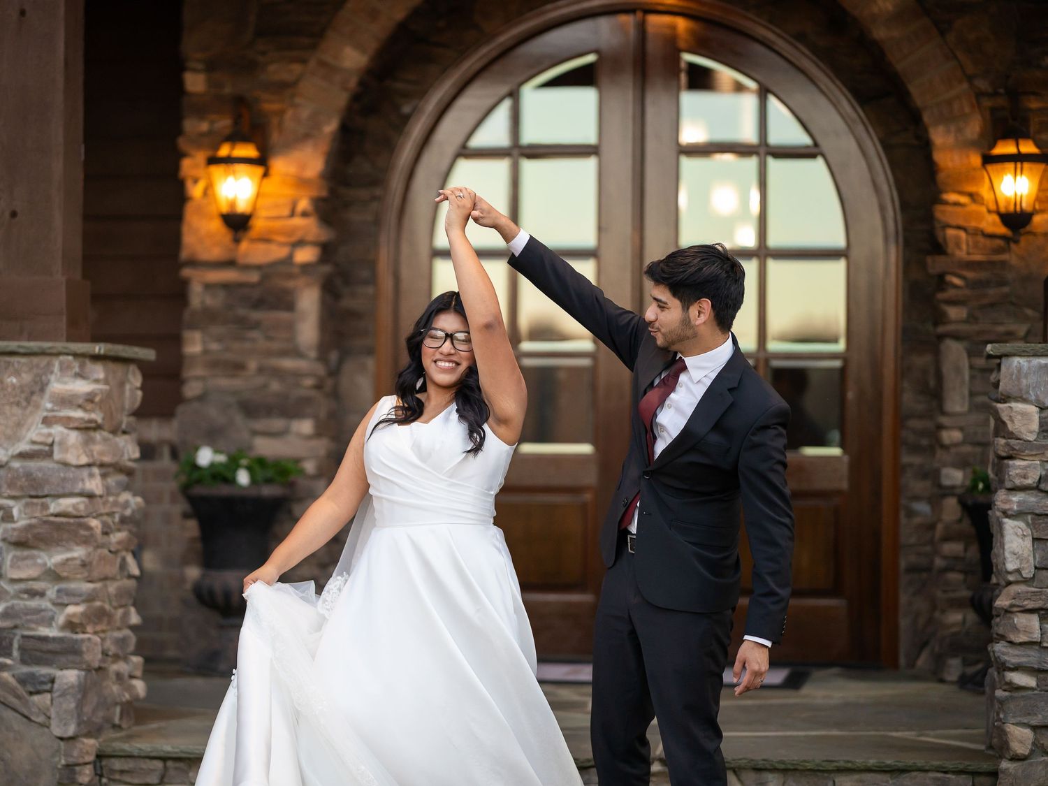 Bride and groom dancing joyfully outside a stone building with arched wooden doors and warm lantern lighting.