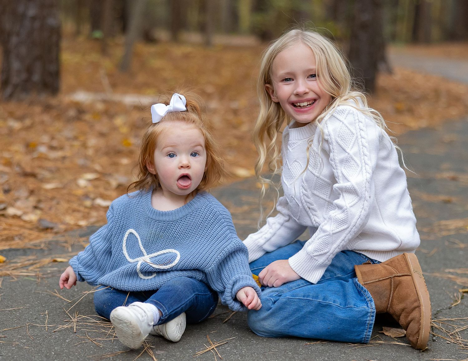 Two children in coordinating blue and white outfits sit together on fallen autumn leaves in an outdoor setting.