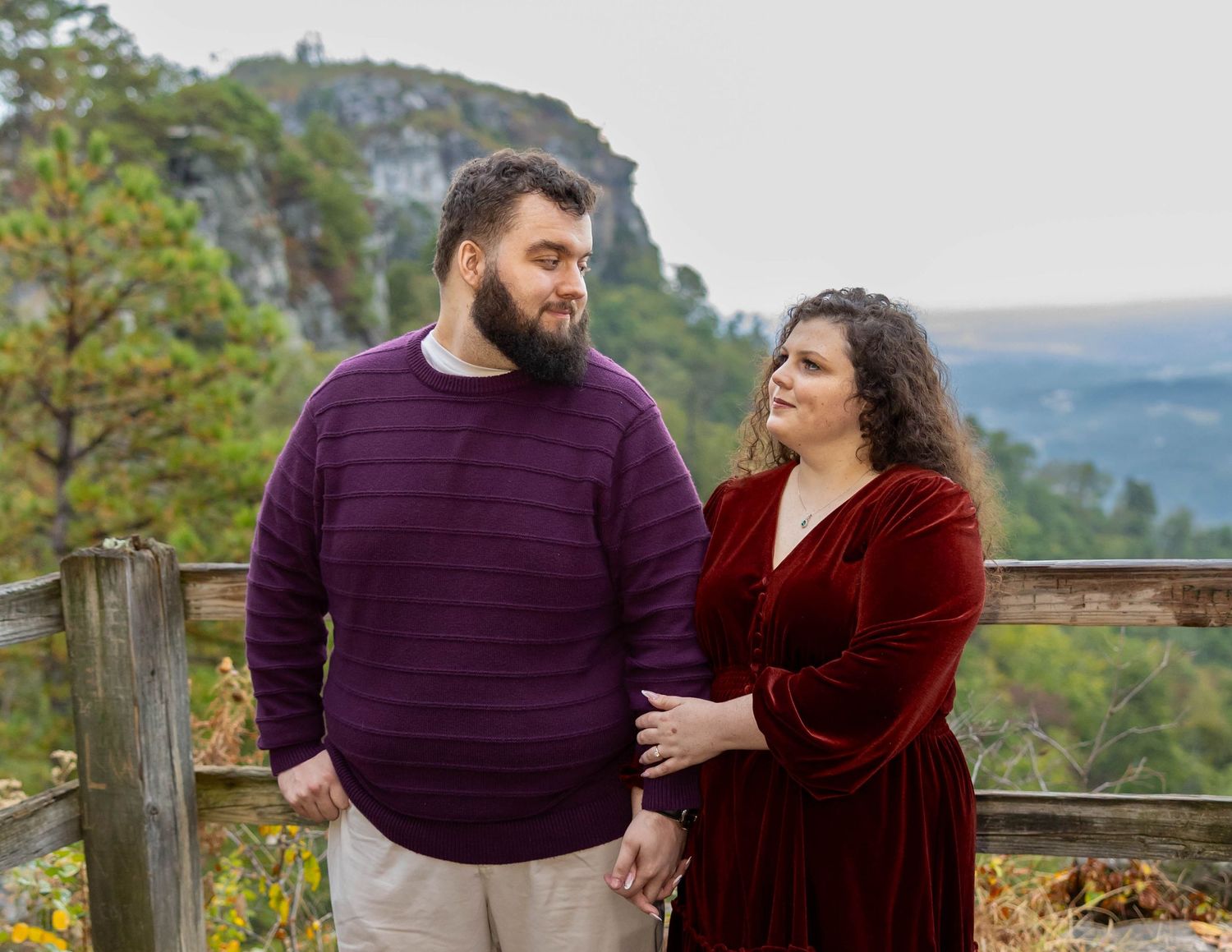 A couple in burgundy and purple sweaters share an intimate moment while standing on a scenic mountain overlook.