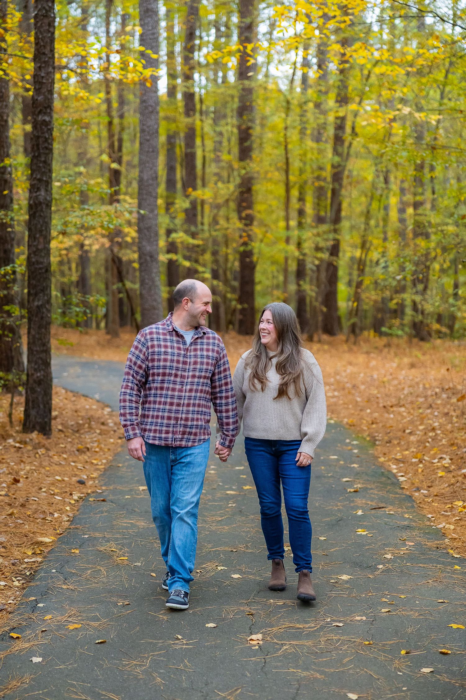 A couple walking hand in hand down a tree-lined path covered in autumn leaves on a fall day.