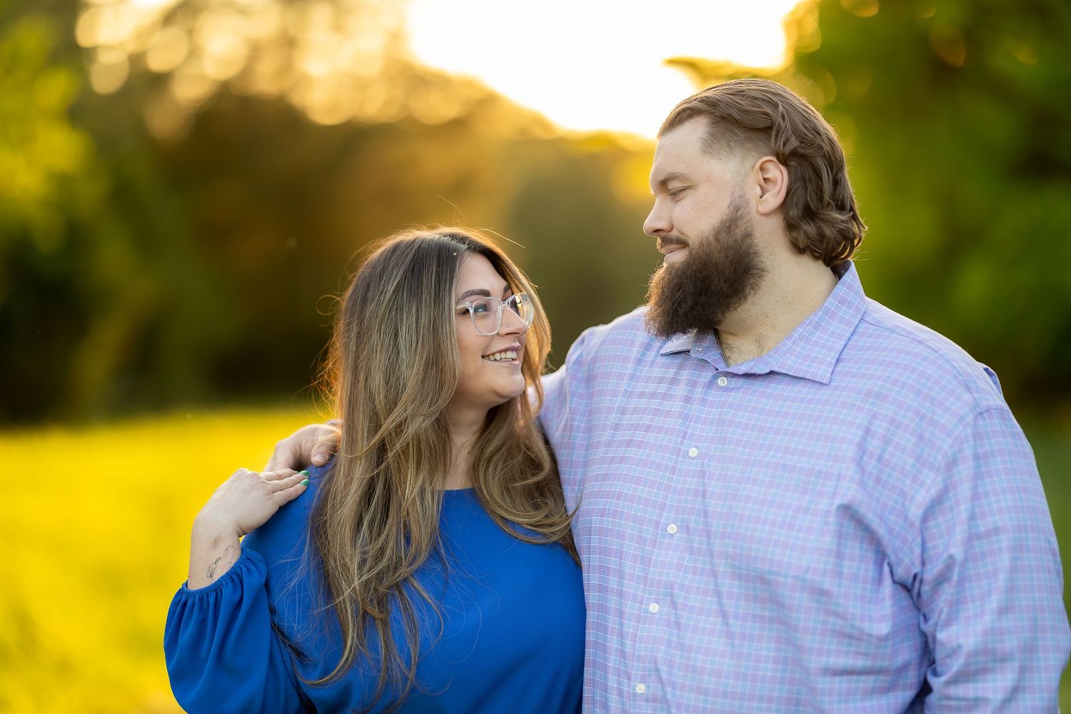 Couple standing together in sunlit field, woman in blue dress, bearded man in plaid shirt, golden hour light.