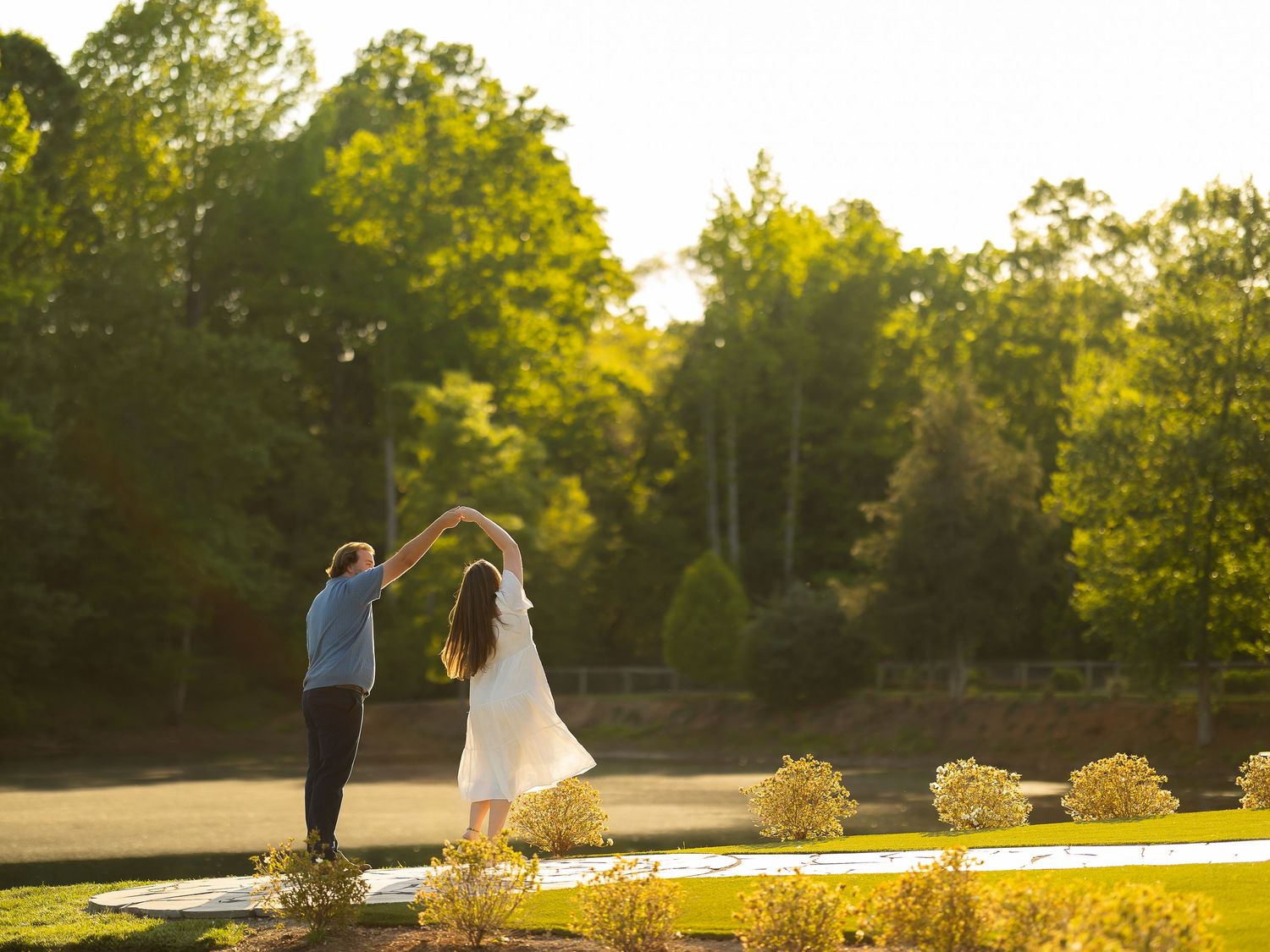 Couple dancing at golden hour in scenic garden with lush green trees and manicured landscape.