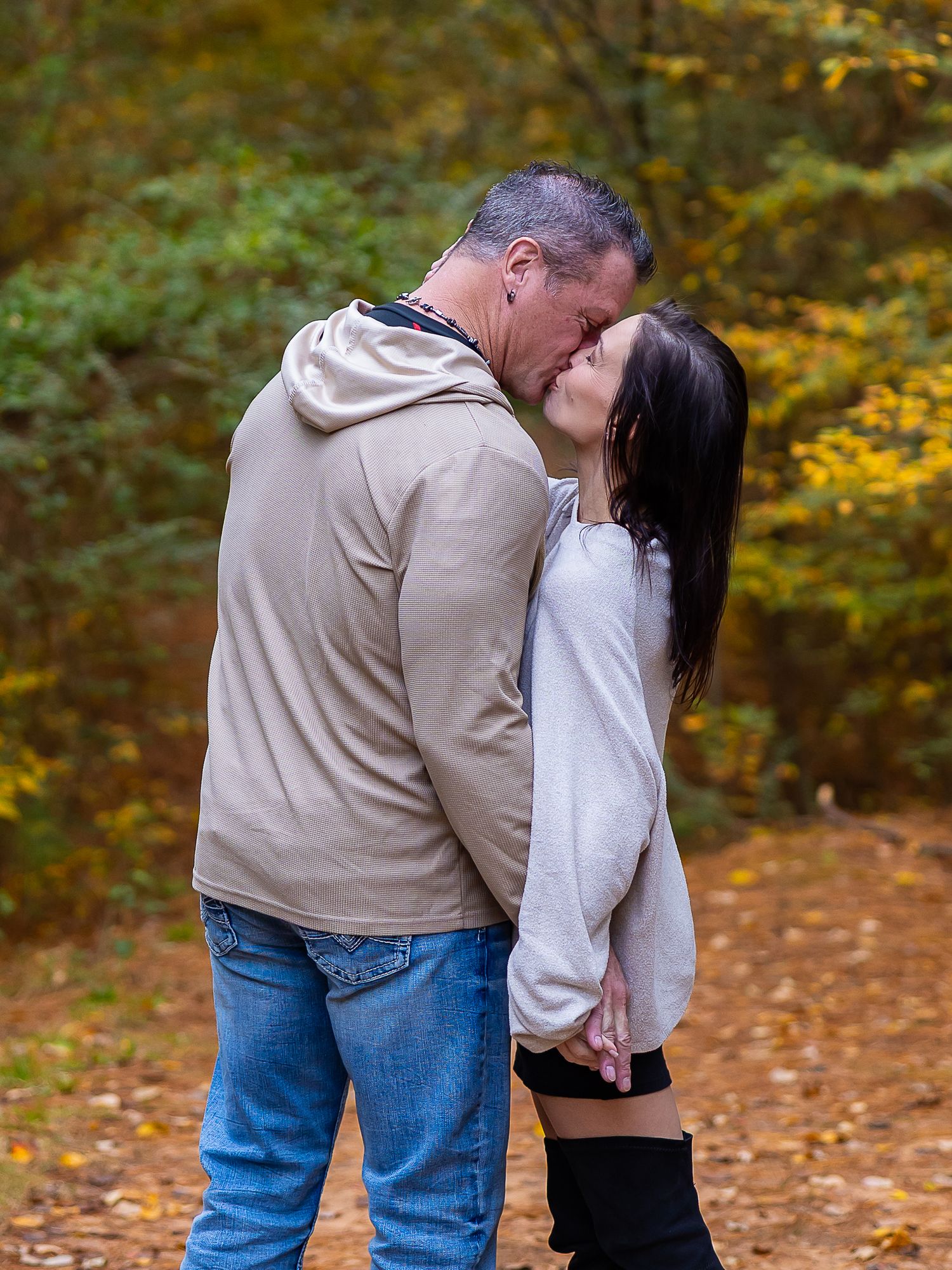 A couple shares a romantic kiss on a path surrounded by autumn foliage in a park setting.