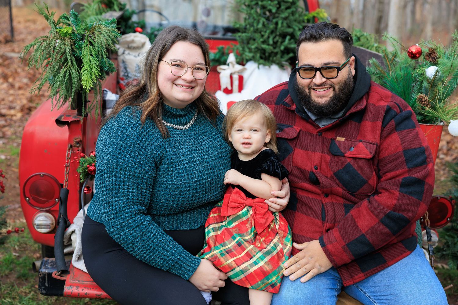 Family poses for Christmas photos outdoors wearing coordinated red and teal outfits with evergreen decorations.