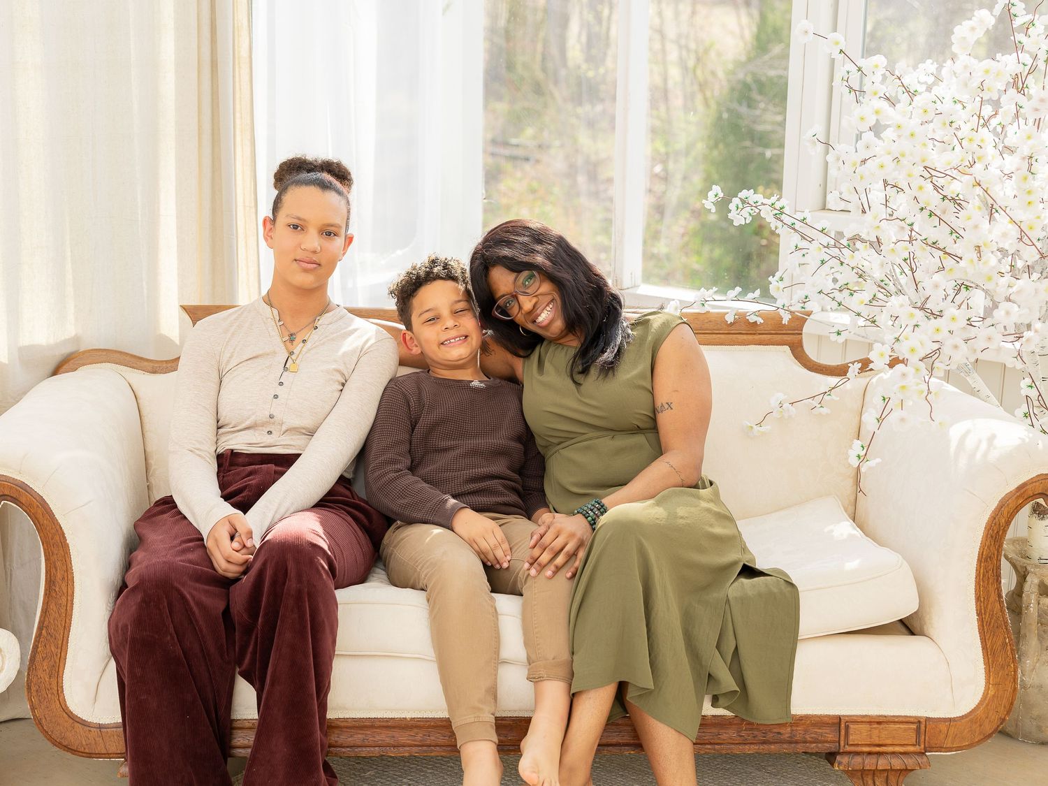 Three women sitting together on a white vintage sofa in a bright room with white floral decor.