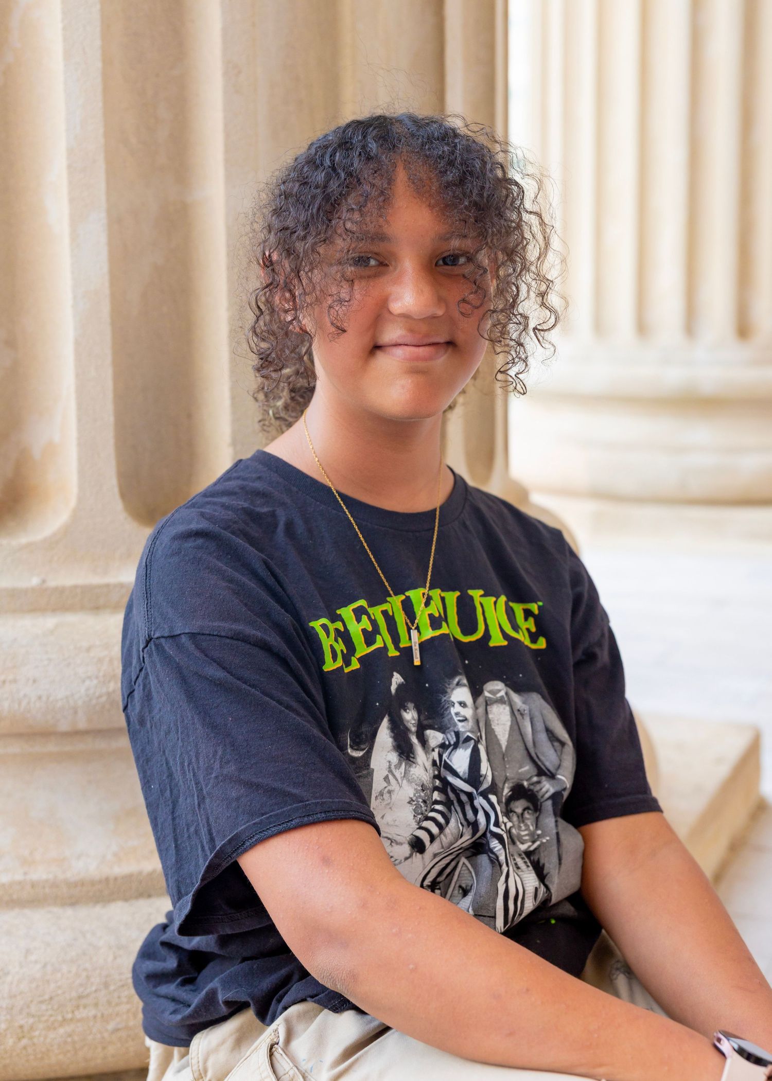 A person wearing a navy Bethune t-shirt with curly hair sits near white pillars in a casual portrait pose.