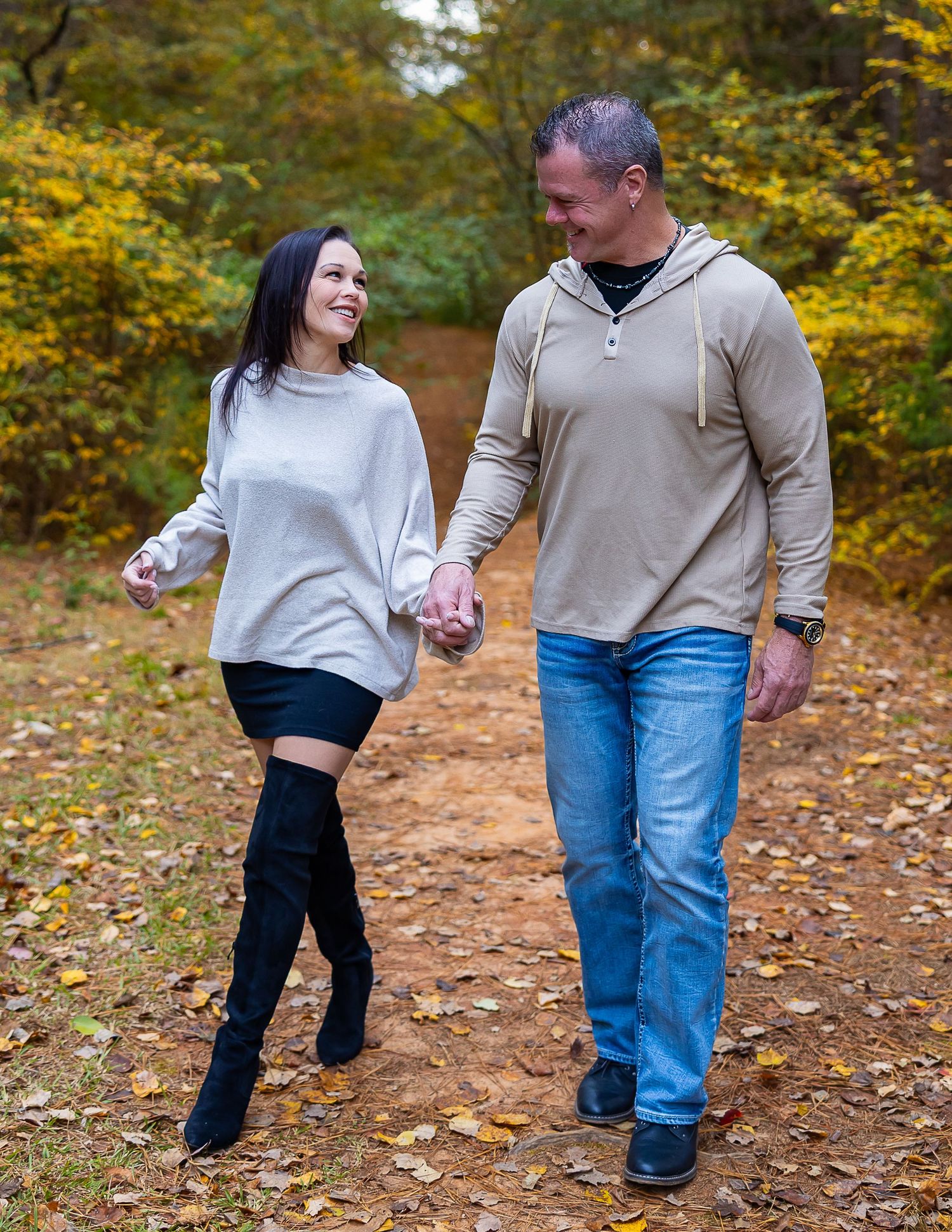 A couple walks hand in hand on a leaf-covered autumn trail through a vibrant forest.