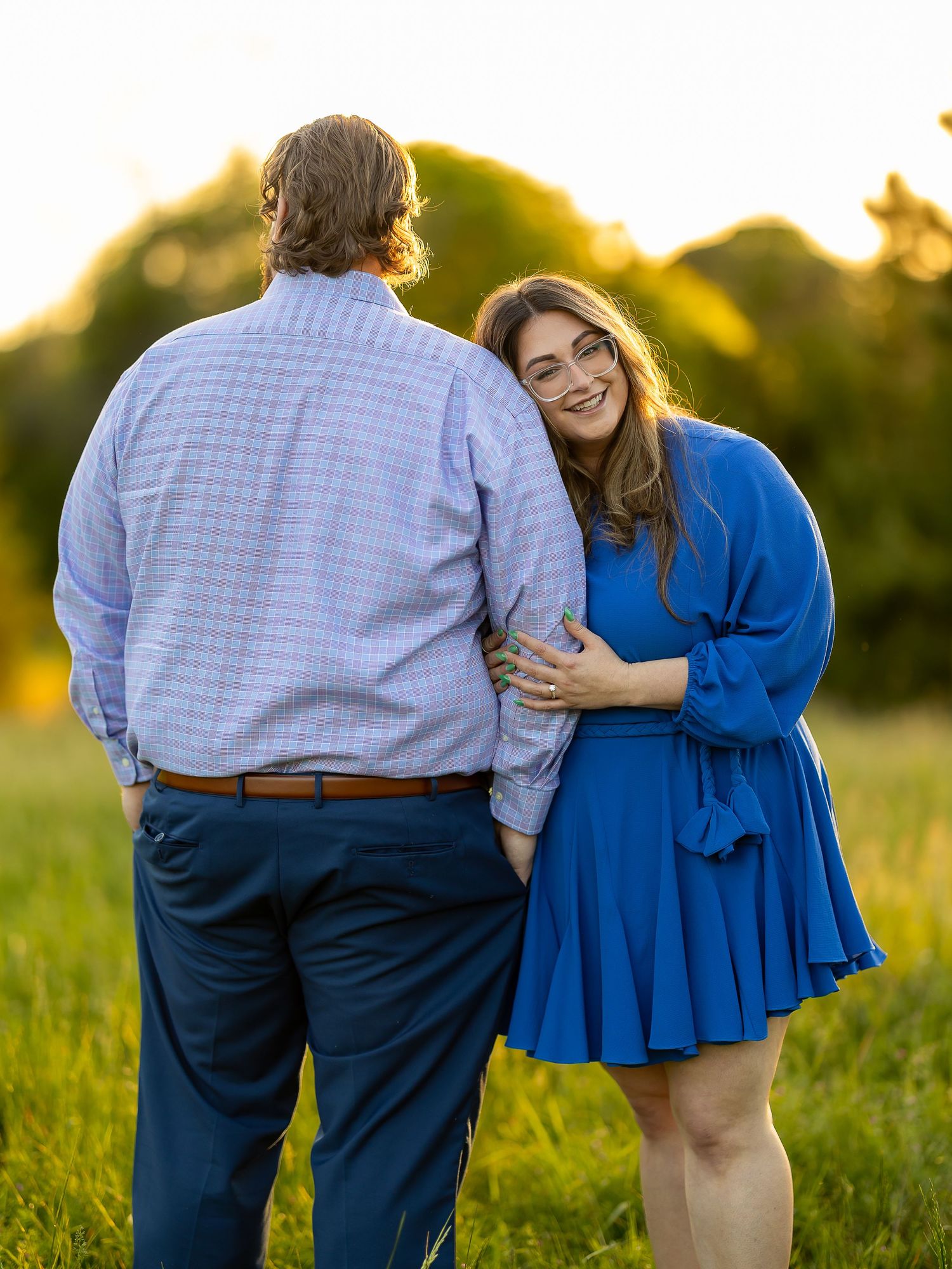 Couple in blue outfits embrace in golden hour field, woman in royal blue dress smiles over shoulder.