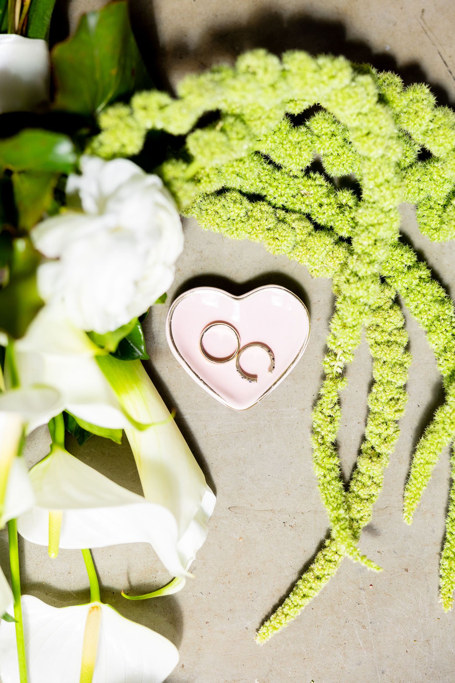 Two gold wedding rings in a pink heart-shaped box surrounded by white calla lilies and green floral accents.