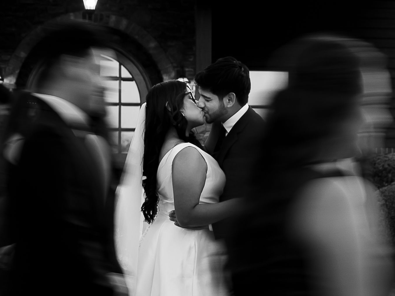 Black and white wedding photo of couple kissing amid blurred guests at elegant venue.