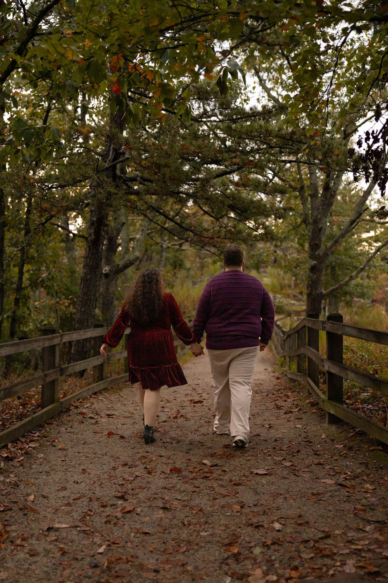 Two people walk together on a wooden boardwalk path through autumn trees with fallen leaves scattered on the ground.