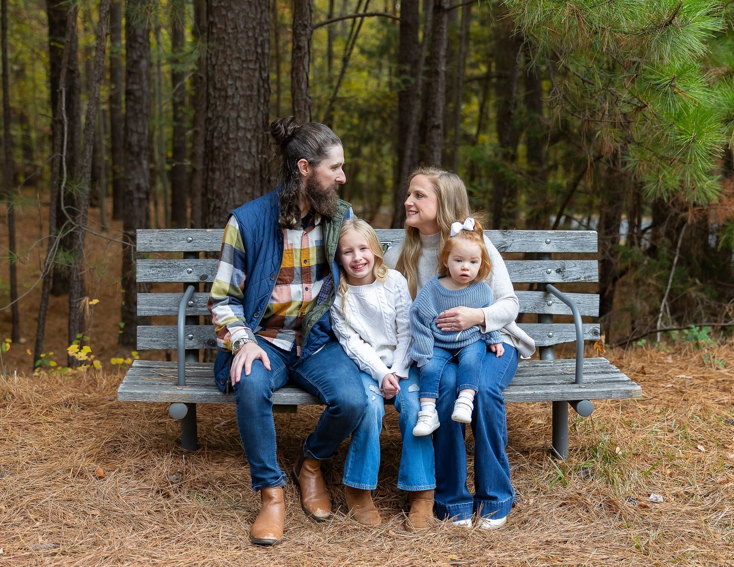 Family members sit together on wooden benches surrounded by tall pine trees in a forest setting.