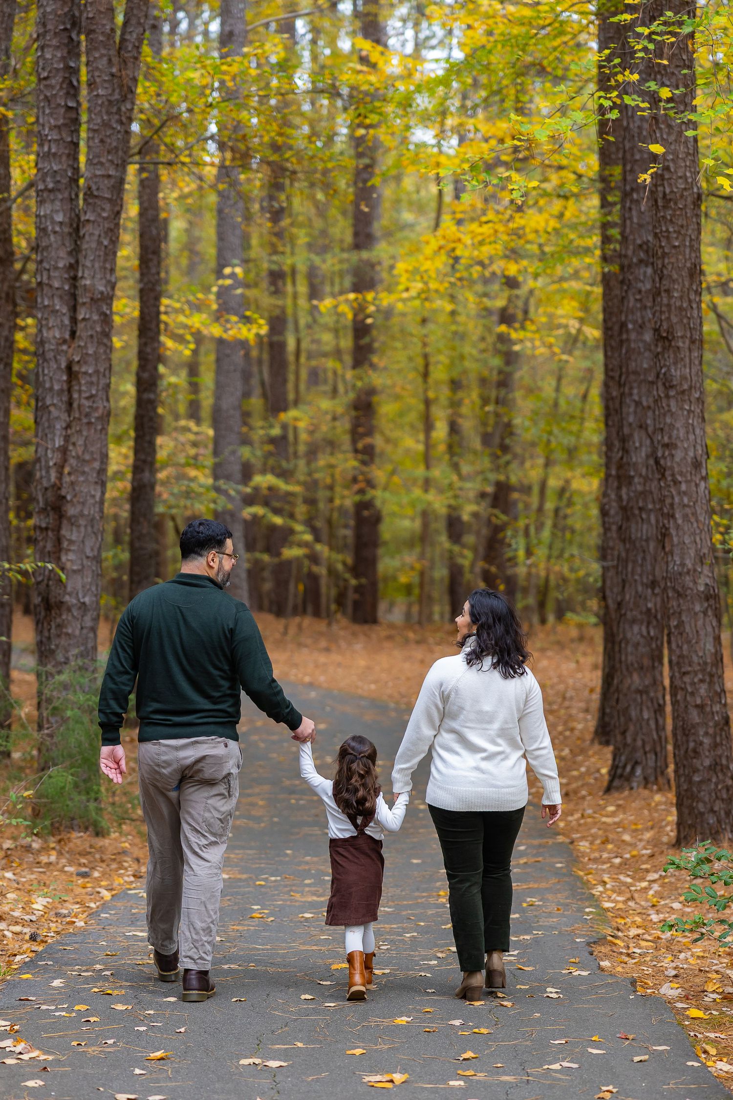 Family walking together on a tree-lined path during autumn with golden yellow foliage and fallen leaves.