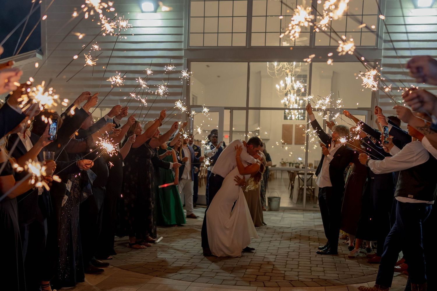 Bride and groom share a romantic kiss during a sparkler send-off at their wedding reception at night.