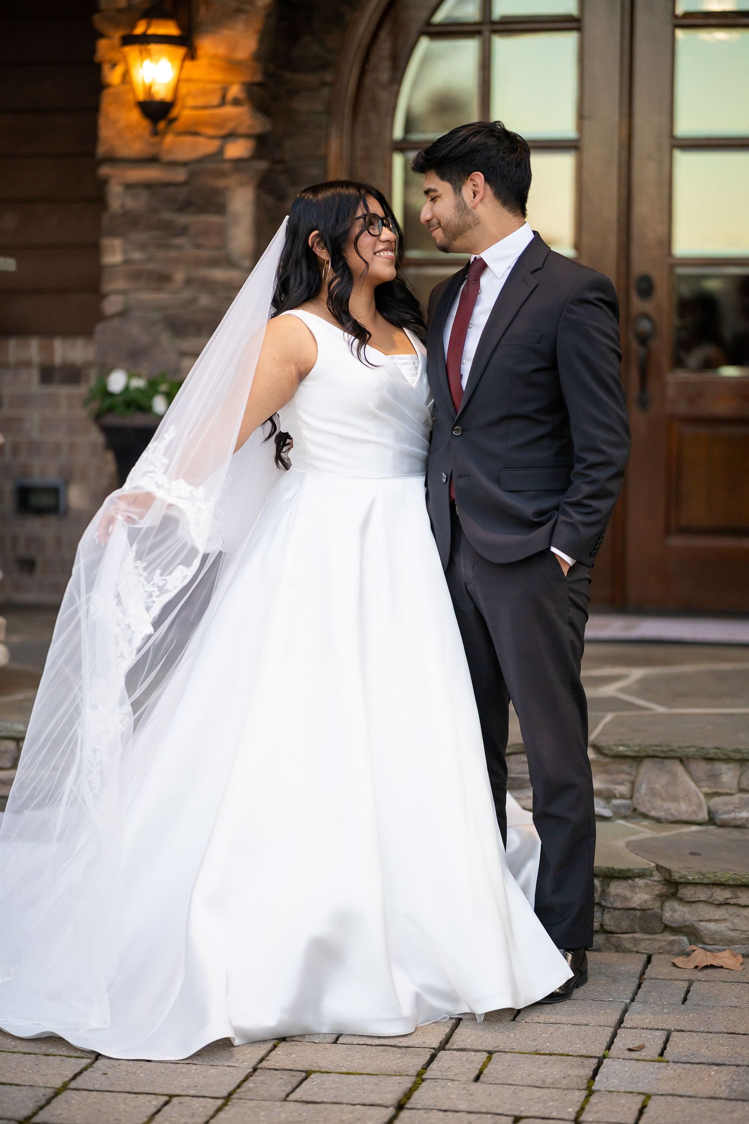 Bride in white gown and veil gazes at groom in gray suit with red tie outside stone venue.
