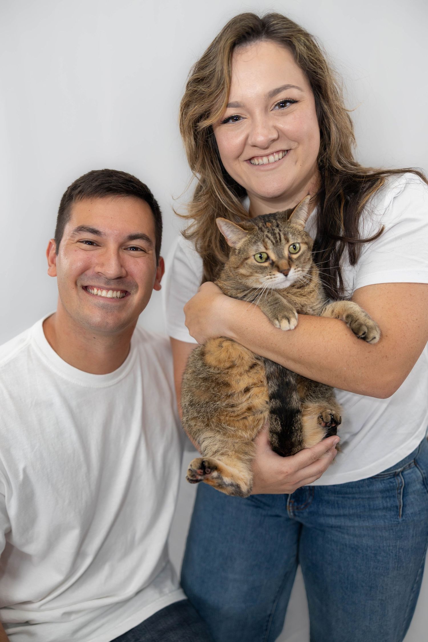 A couple poses with their expressive tabby cat in a series of playful studio portraits against a light background.