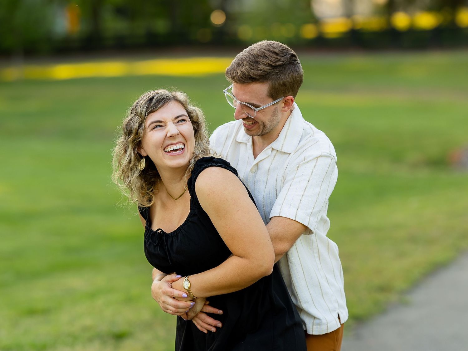 A laughing couple embraces outdoors in a park, woman in black dress and man in white shirt, sharing a joyful moment.