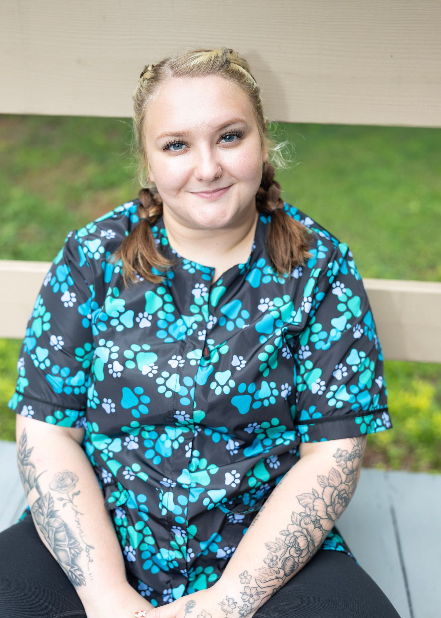 Healthcare worker in paw print scrubs and tattoos sitting outdoors on a sunny day.