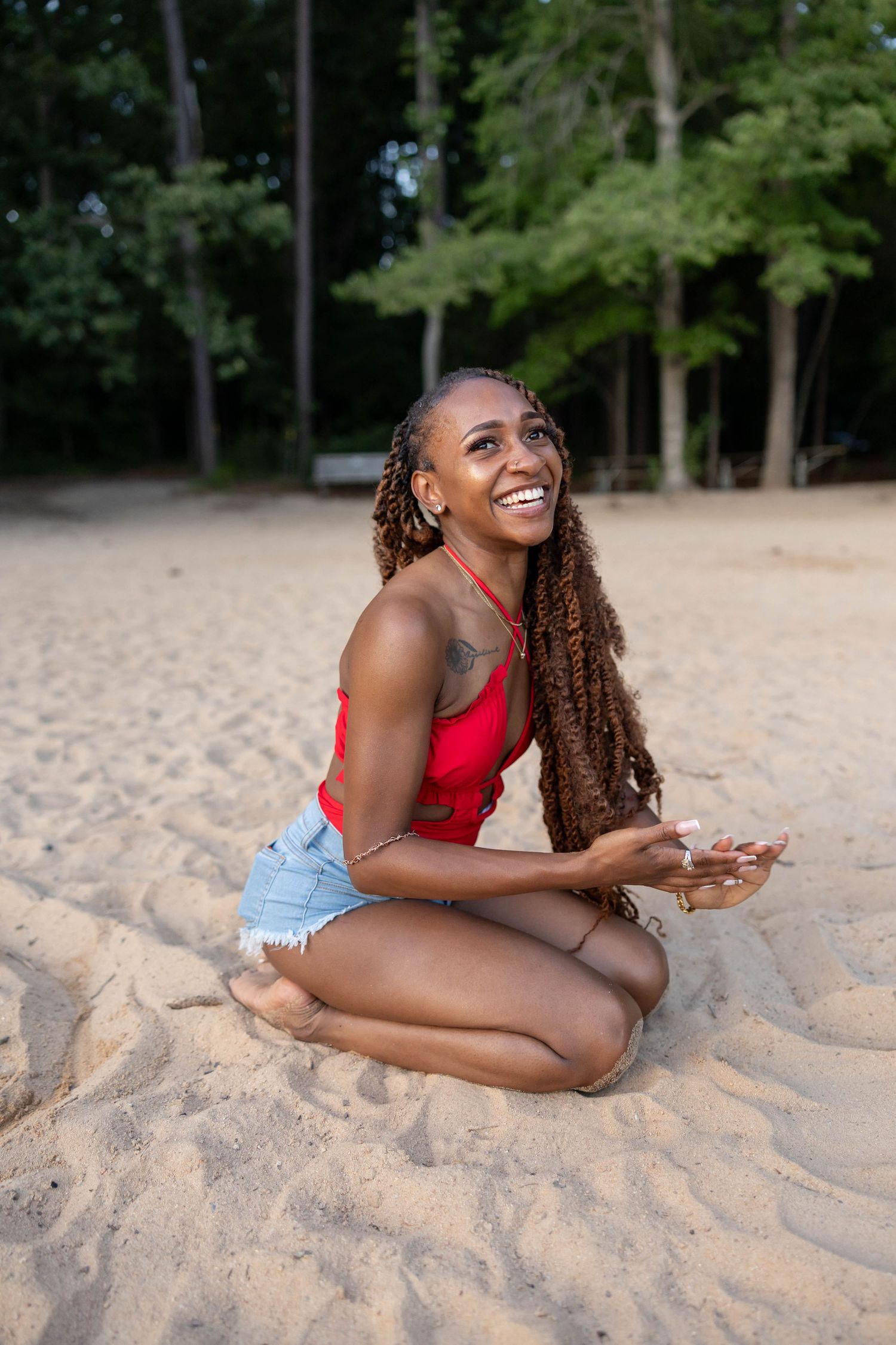 Woman with long braided hair wearing red tank top and denim shorts sits laughing on sandy beach near trees.