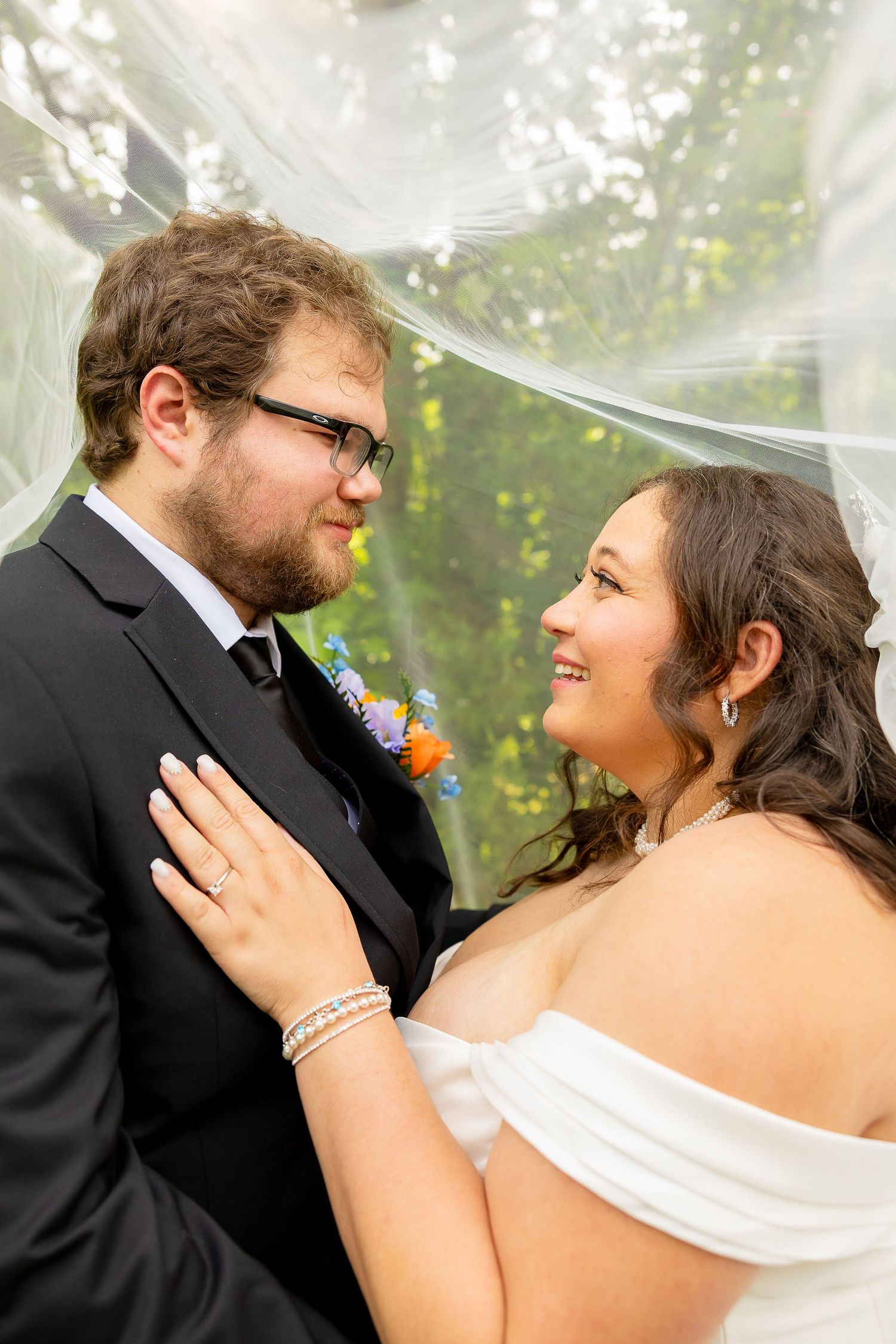 Bride and groom share a tender moment under a flowing veil outdoors on their wedding day.