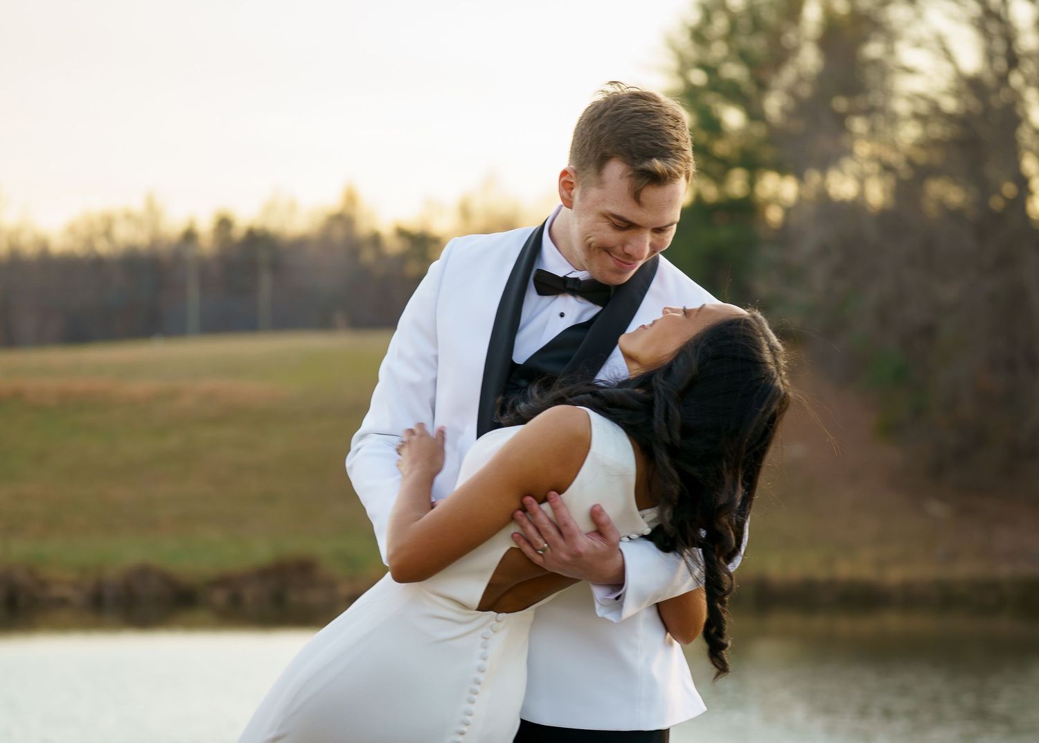 Bride and groom share a romantic dip pose near a serene pond at golden hour during their outdoor wedding.