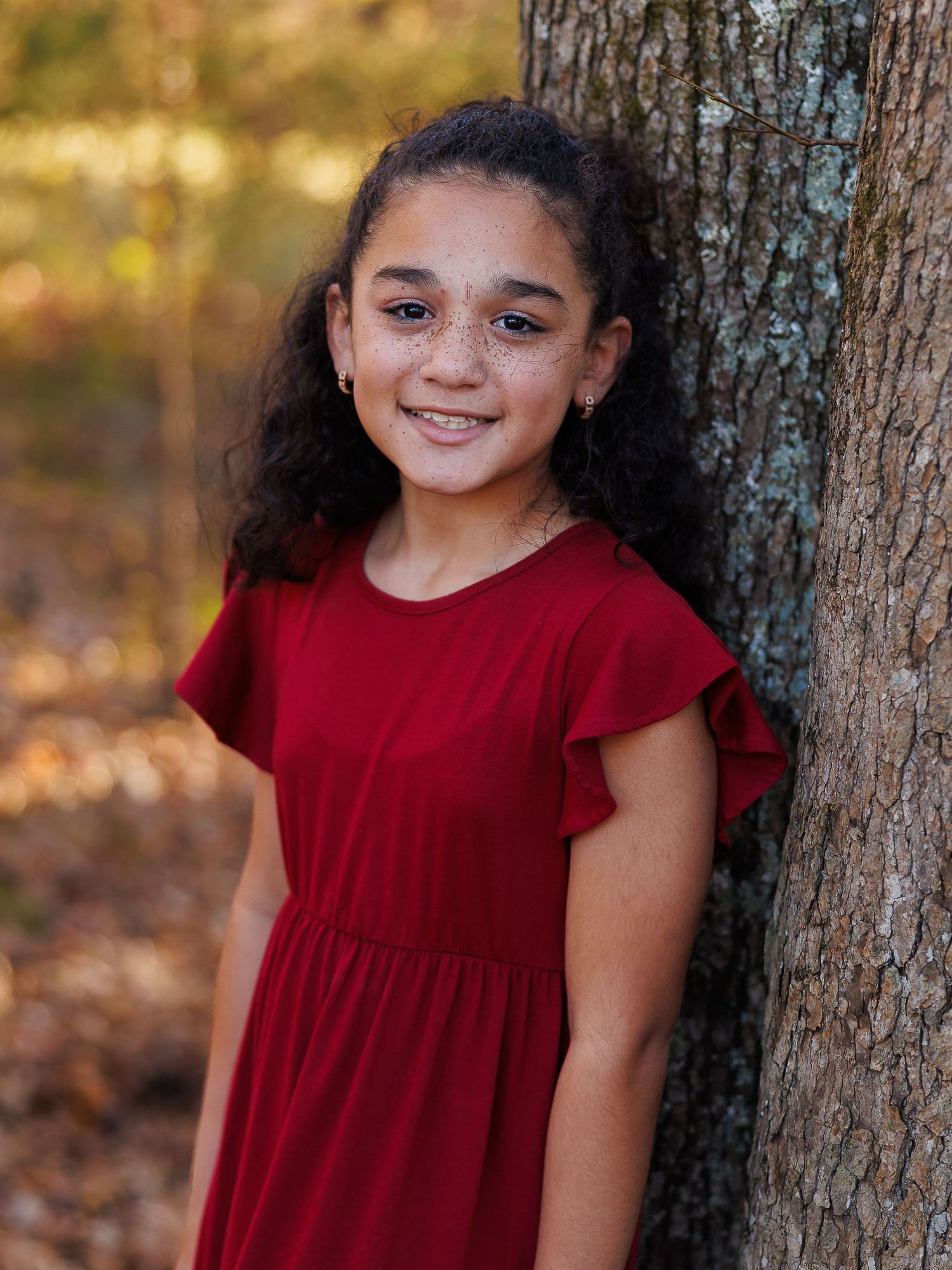 A child in a red dress poses near a tree trunk in an autumn forest setting with warm natural lighting.