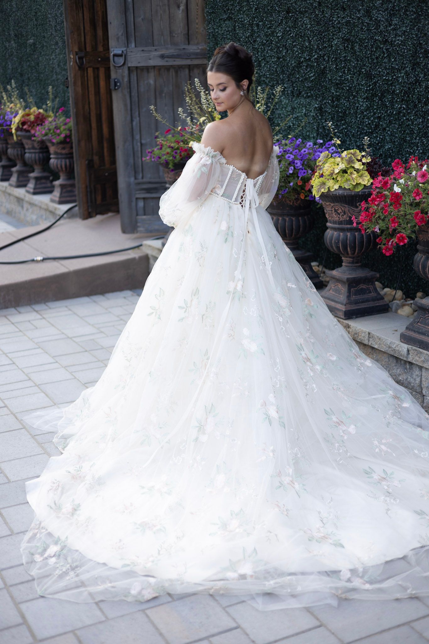 A stunning off-shoulder wedding gown with delicate lace details and long train photographed against colorful garden flowers.