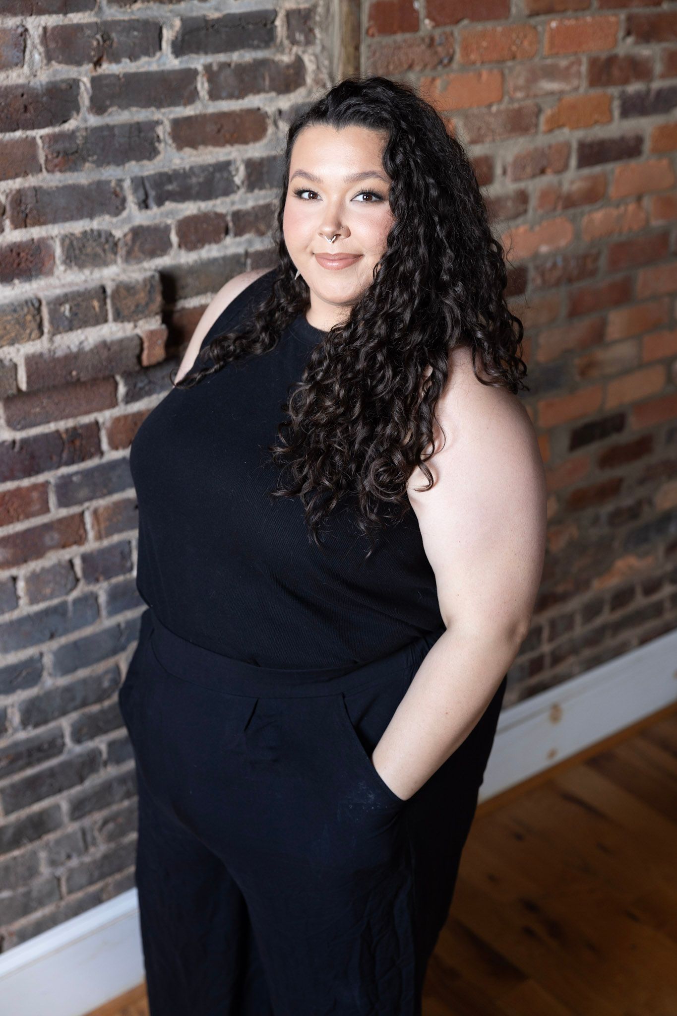 Professional portrait of a person in a black sleeveless outfit posing against a brick wall background.