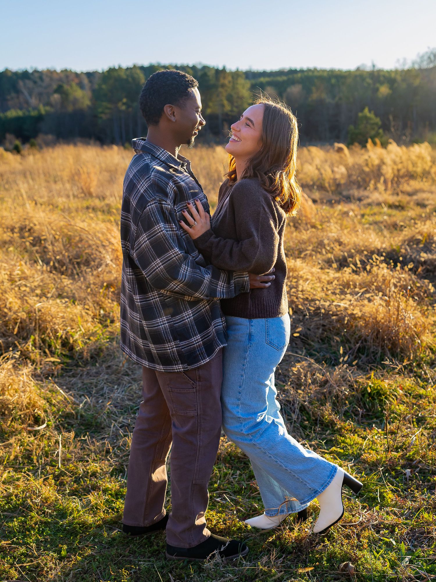 A couple embraces in a sunlit meadow during golden hour with trees in the background.