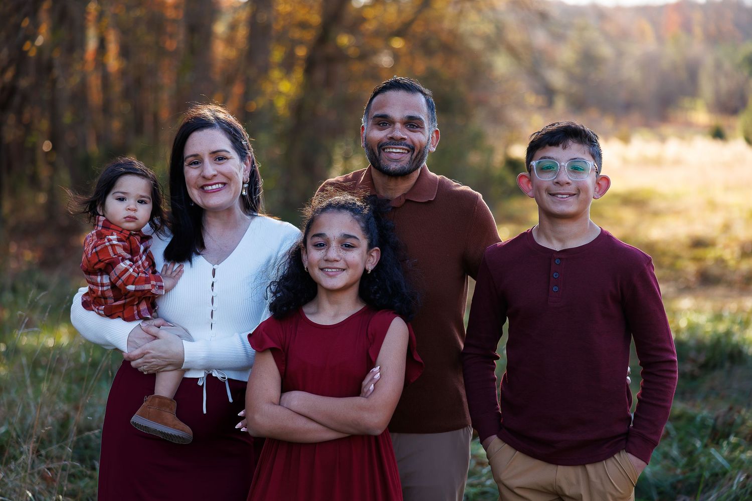 A family in coordinating burgundy and white outfits poses together in a wooded setting during fall.