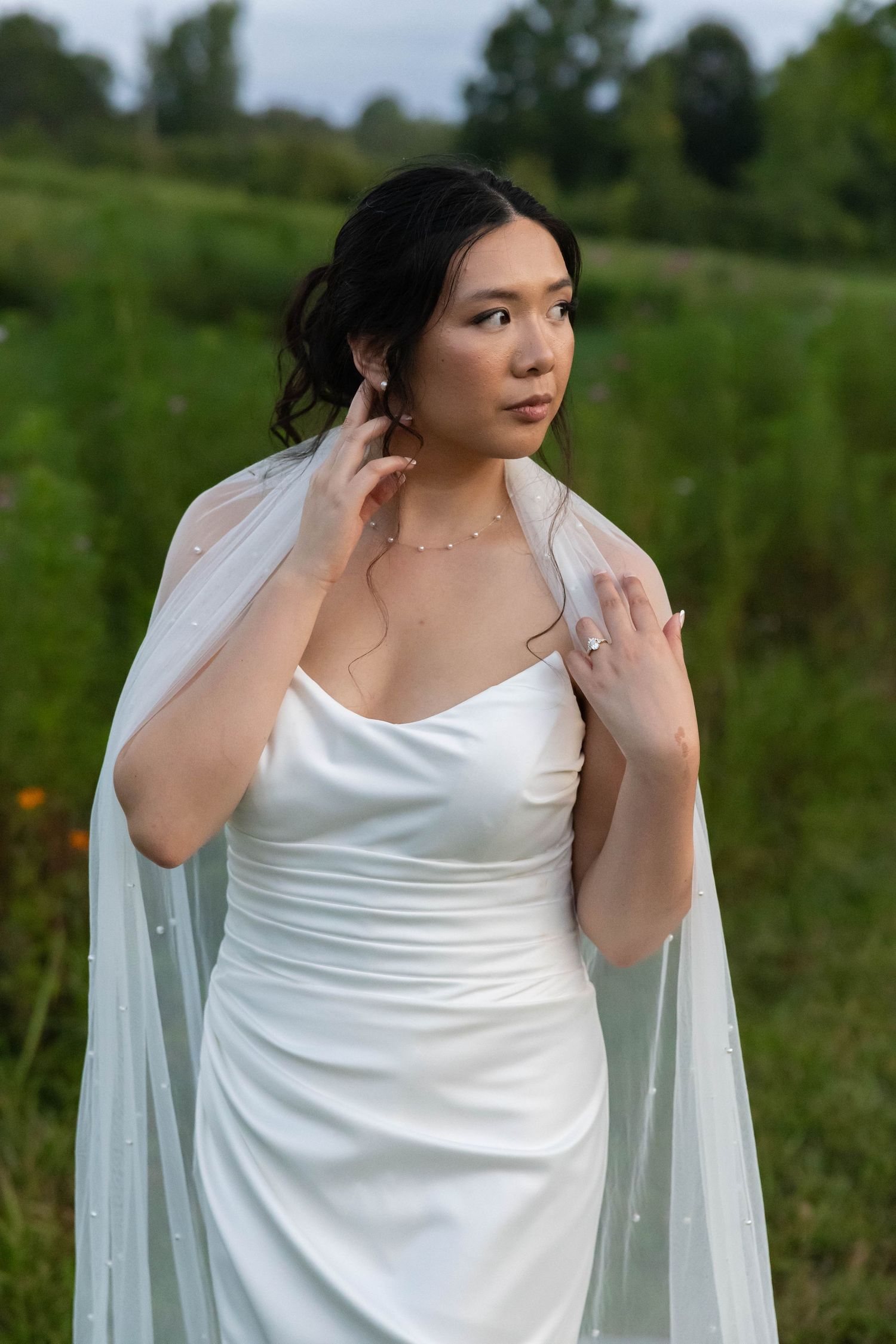 A bride in a white dress adjusts her flowing veil while posing outdoors in a natural green setting.