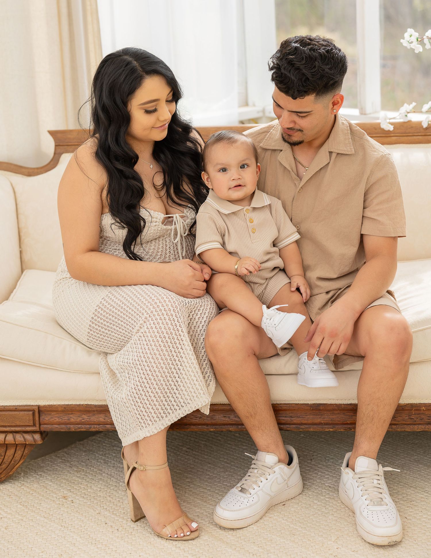 Family sitting together in coordinated neutral outfits on wooden bench in bright sunlit room with white walls.