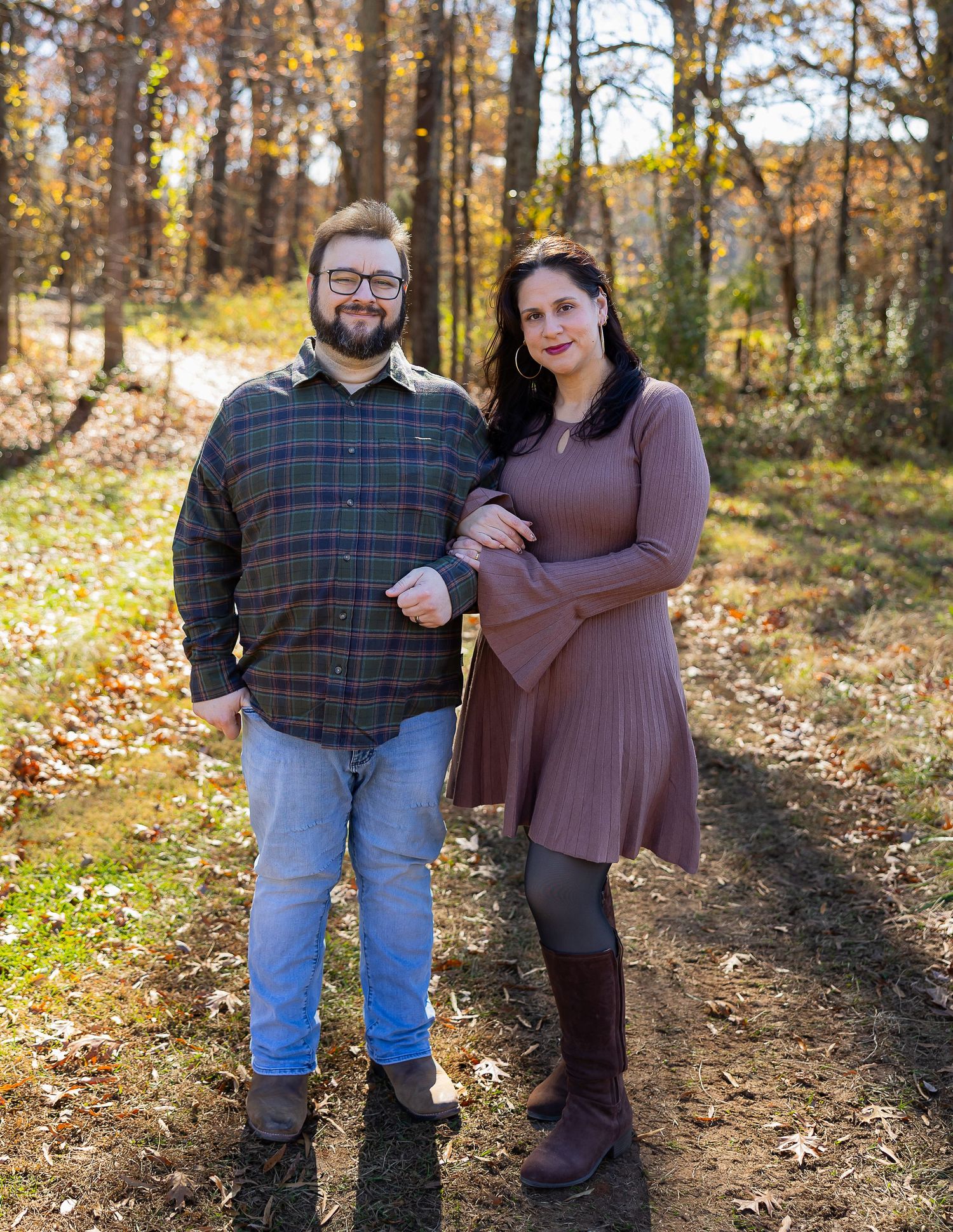 A couple stands together on a leaf-covered forest path during autumn with warm golden sunlight filtering through trees.