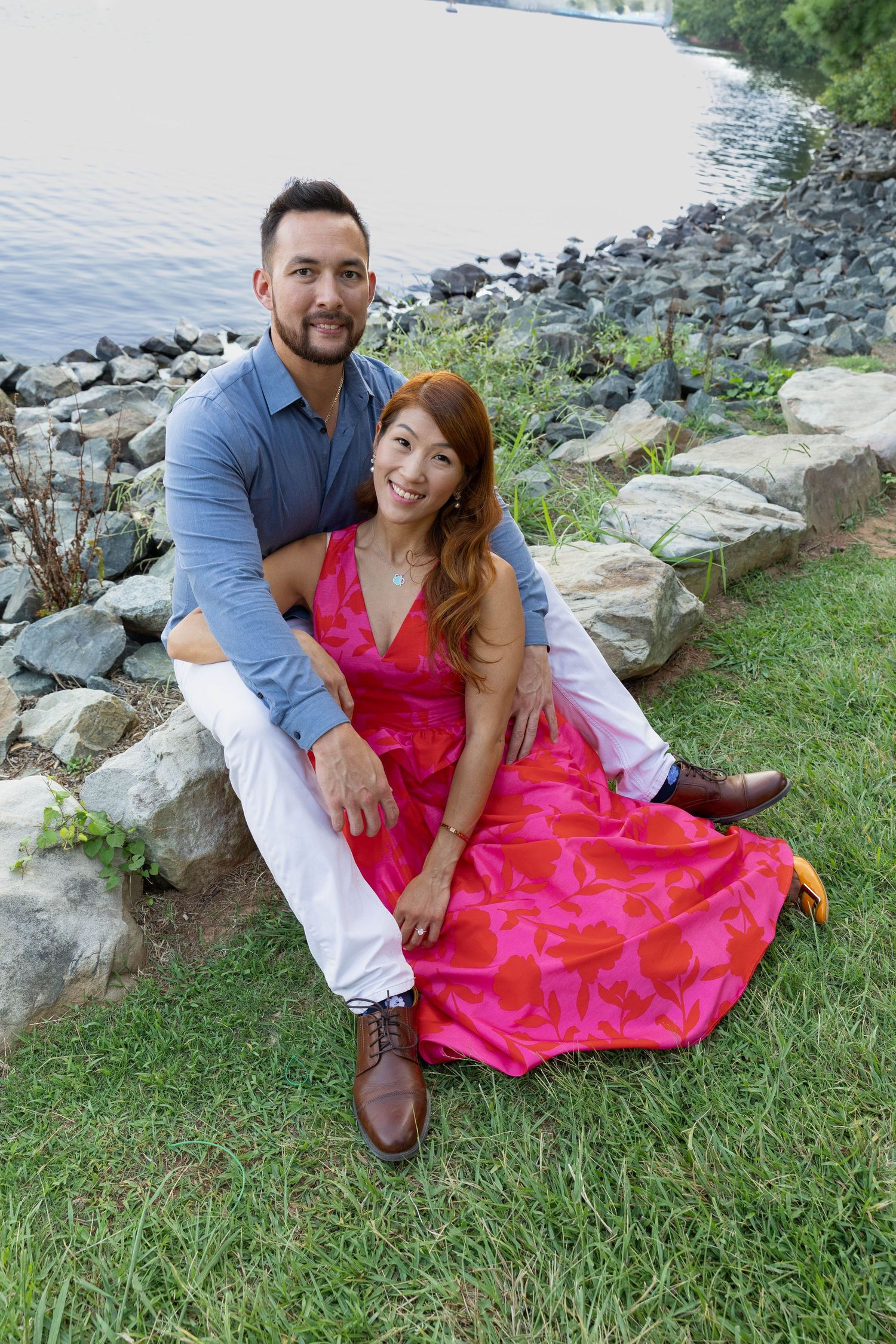A couple in blue and pink attire poses together on grassy ground near a rocky shoreline on a sunny afternoon.