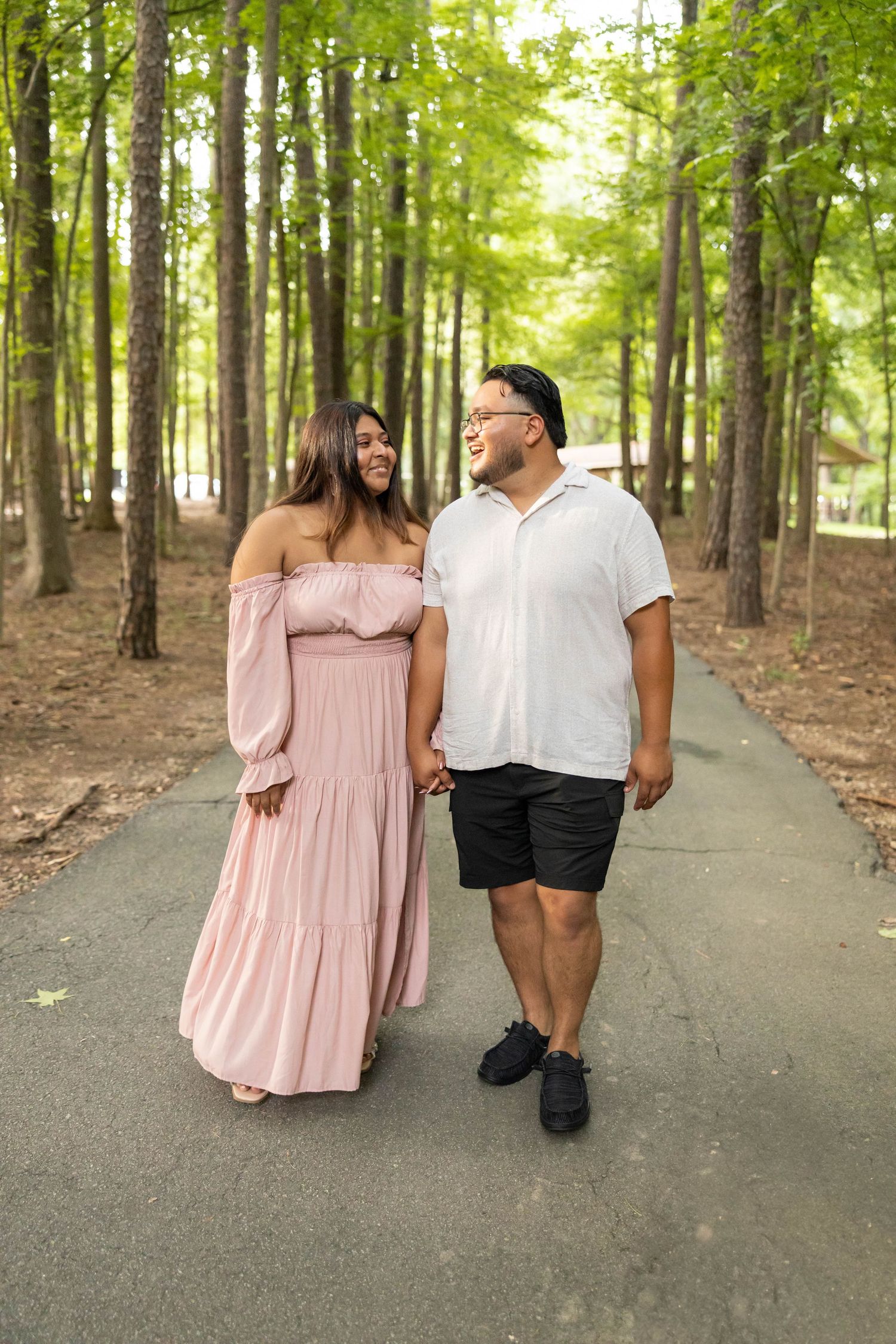 A couple walks together on a paved path through a lush green forest on a sunny day.