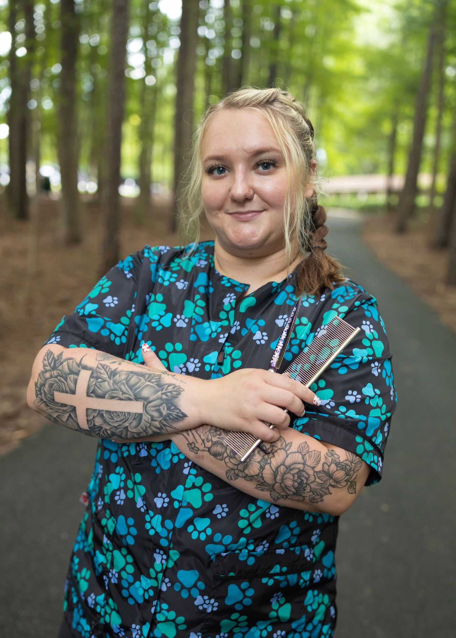 Healthcare worker in pawprint scrubs with tattoos holding knitting needles stands in a wooded forest setting.