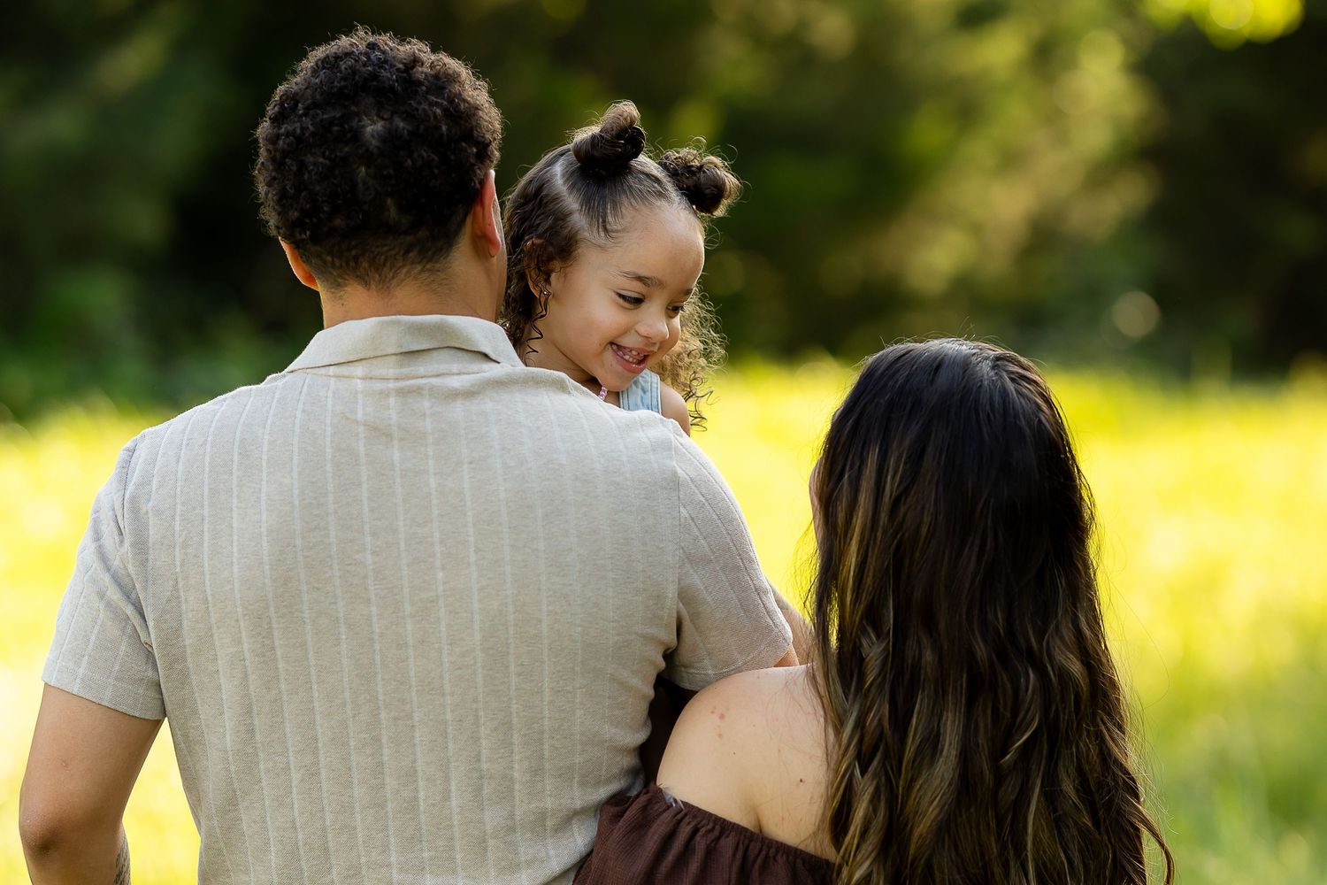 A family of three enjoys a sunny day outdoors, with parents holding their young daughter in a green field.