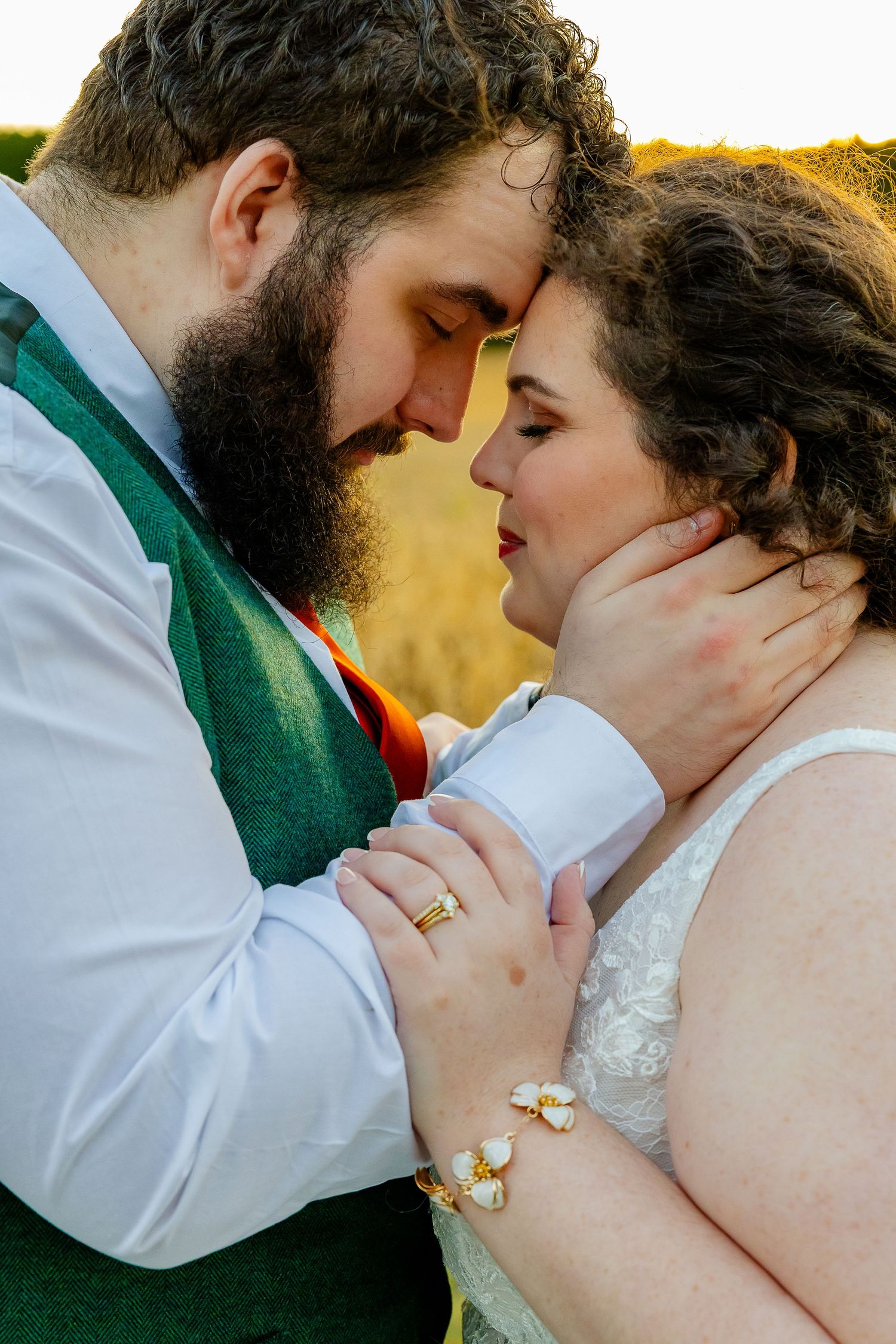 Groom gently cupping brides face as they share an intimate moment, bride wearing gold floral bracelet at sunset.
