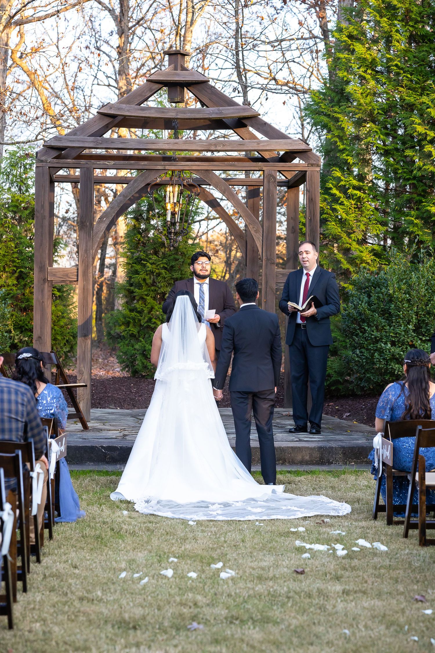 Outdoor wedding ceremony under rustic wooden gazebo with bride in white gown and veil facing officiant.