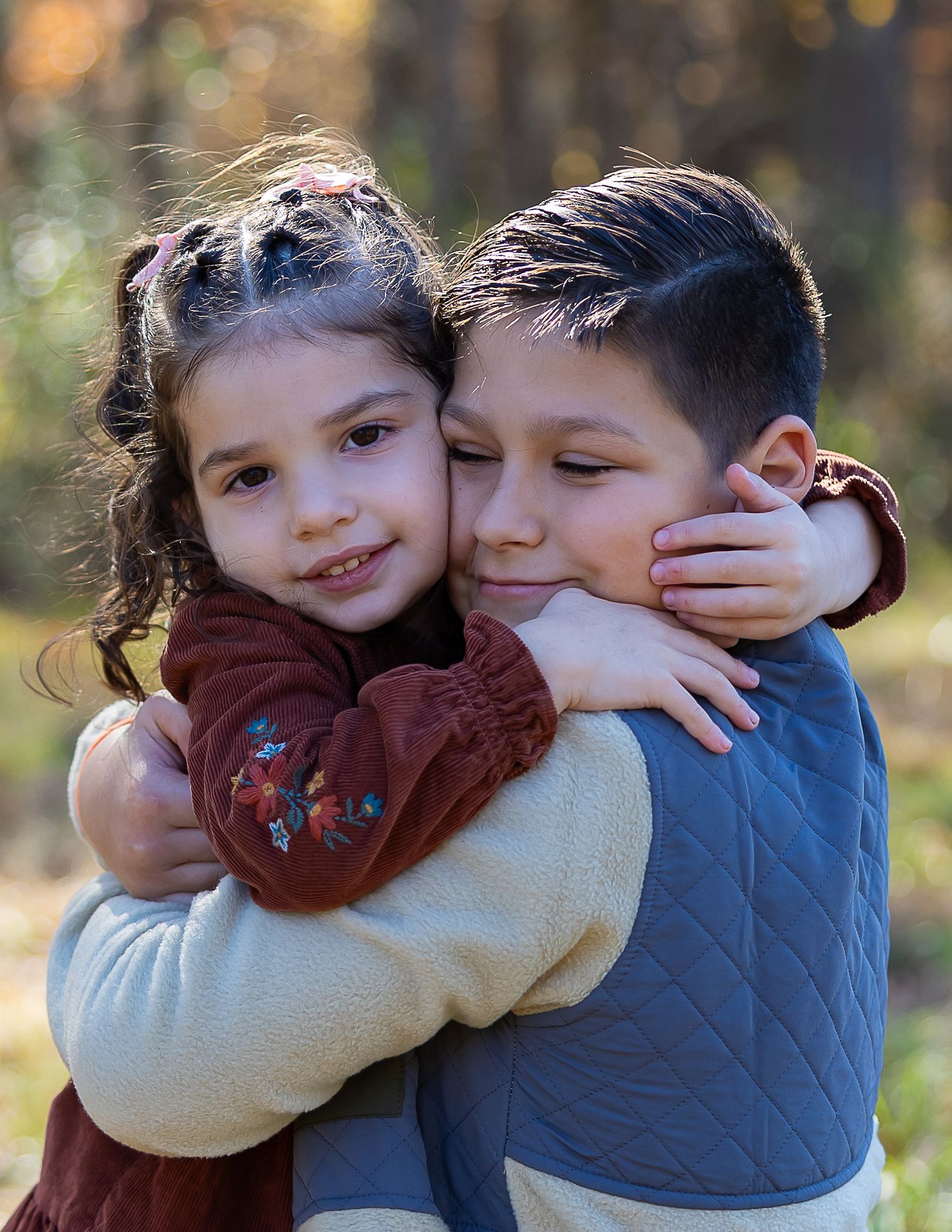 Two children share a warm embrace outdoors with autumn trees in the blurred background.