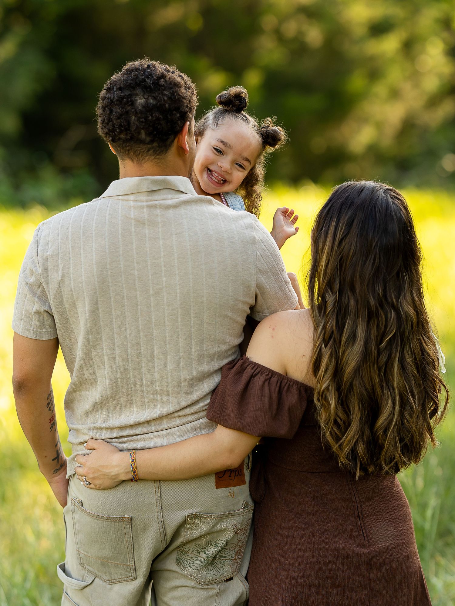 A family of three poses outdoors, parents holding their laughing toddler girl in a sunny green field.