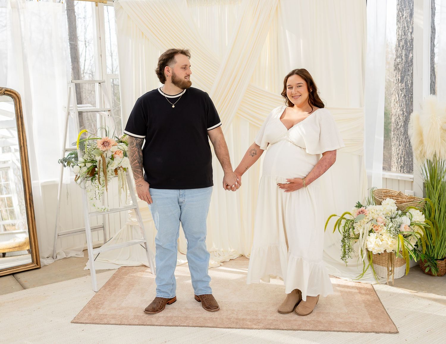 A couple holds hands while standing together in a bright, airy room with white decor and indoor plants.