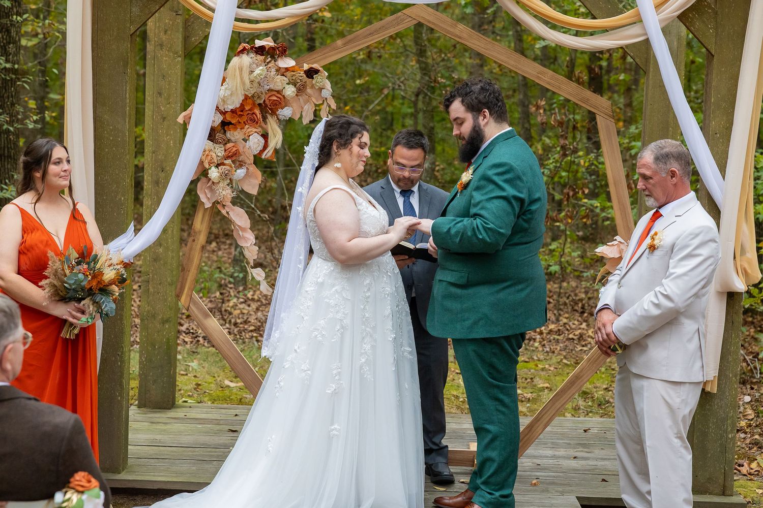Couple exchanging vows at outdoor fall wedding ceremony under wooden arch with white draping and autumn floral decor.