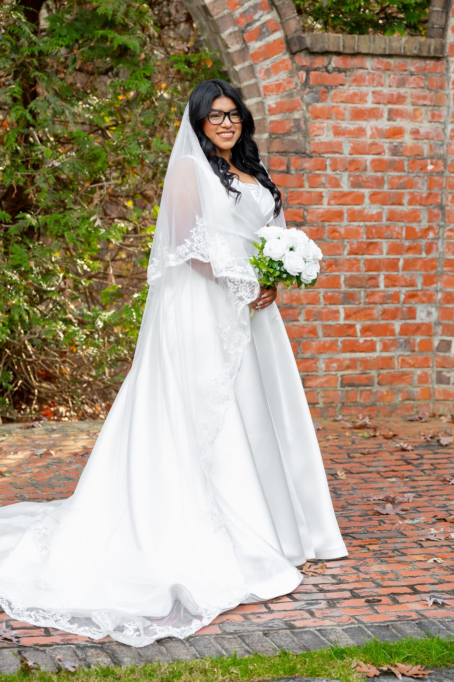 Smiling bride in white gown and veil holding bouquet beside brick wall outdoors.