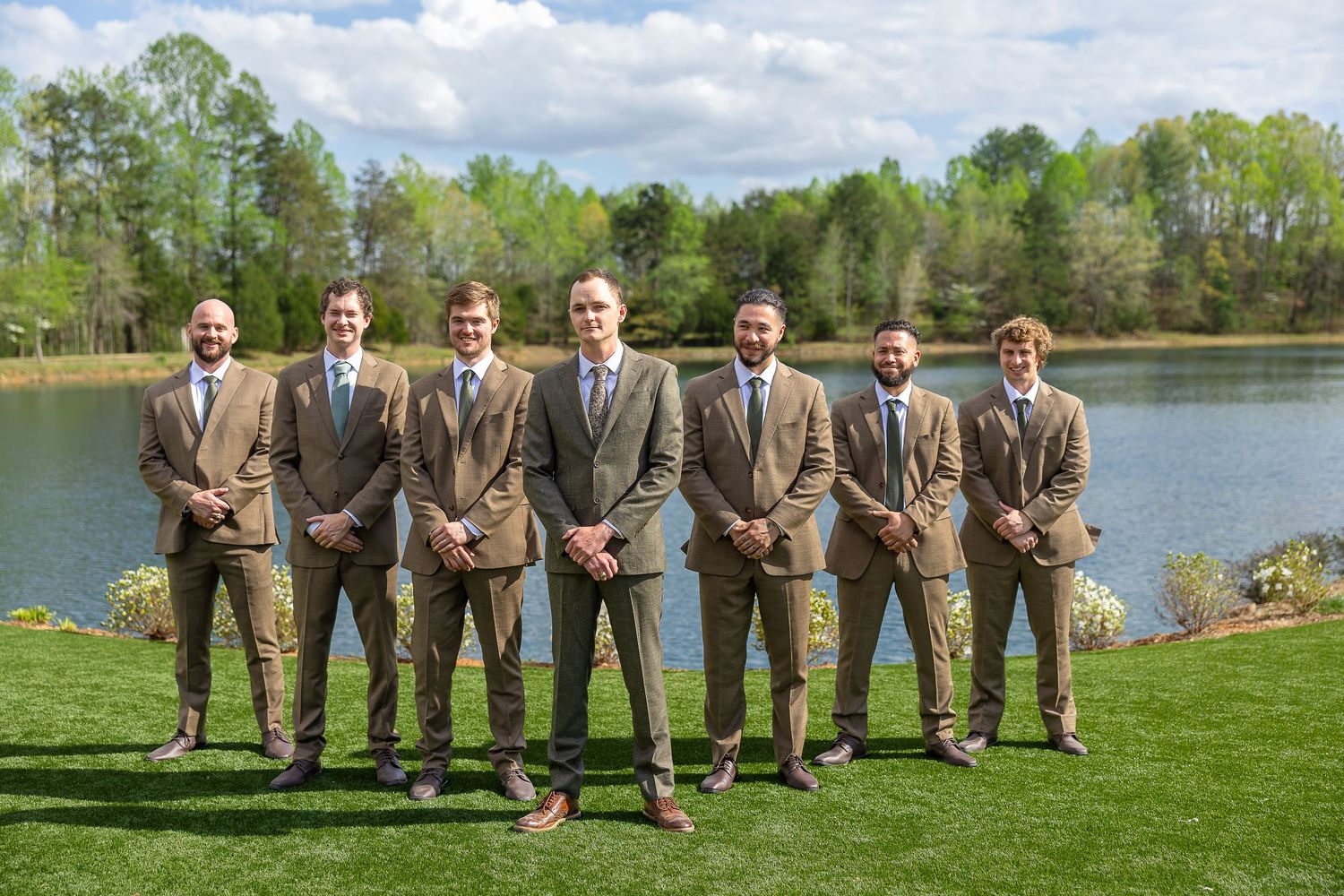 Six groomsmen in matching brown suits pose outdoors by a serene lake surrounded by lush green trees on a wedding day.