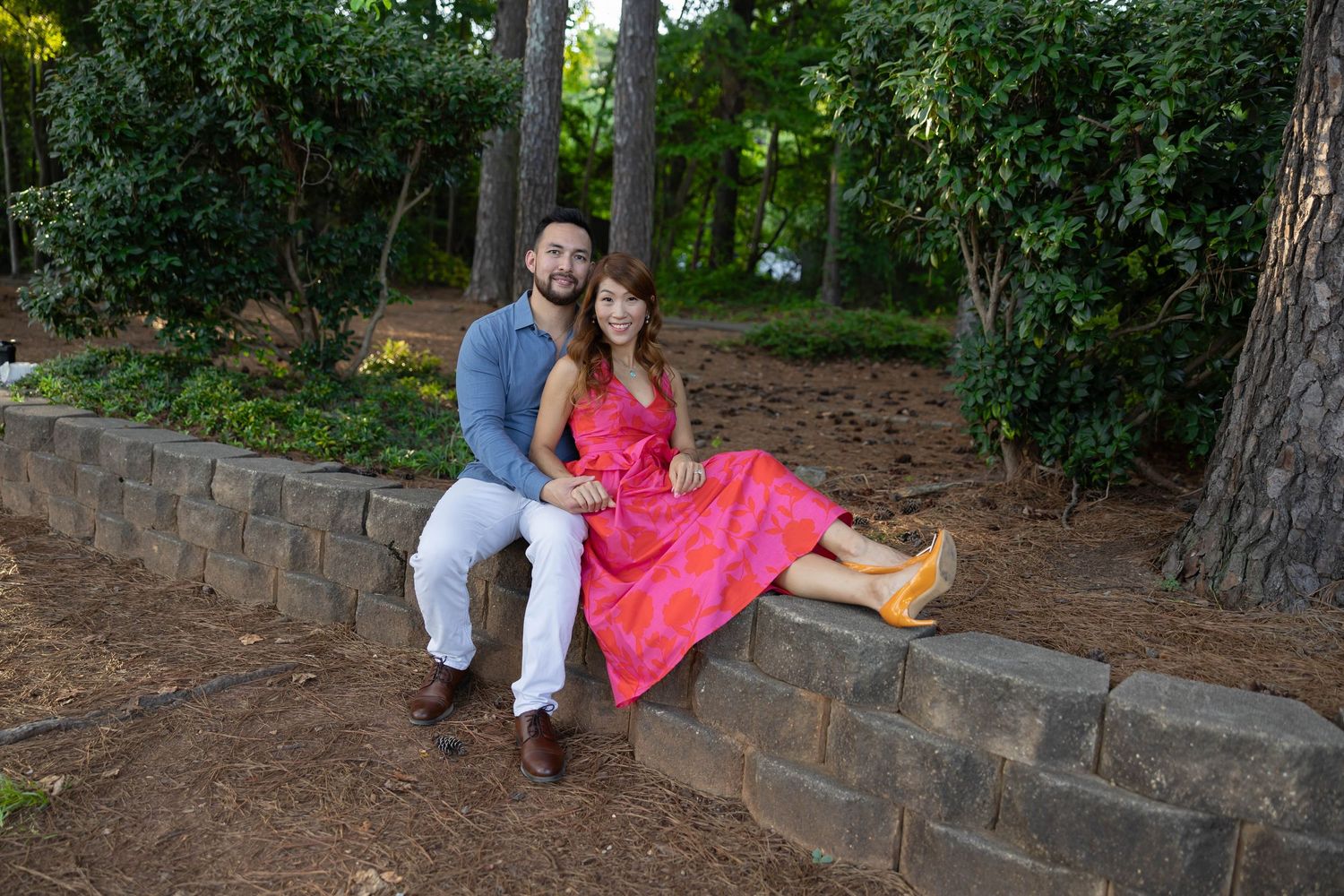 A couple sits together on a stone retaining wall in a garden setting at dusk wearing coordinated pink and blue outfits.