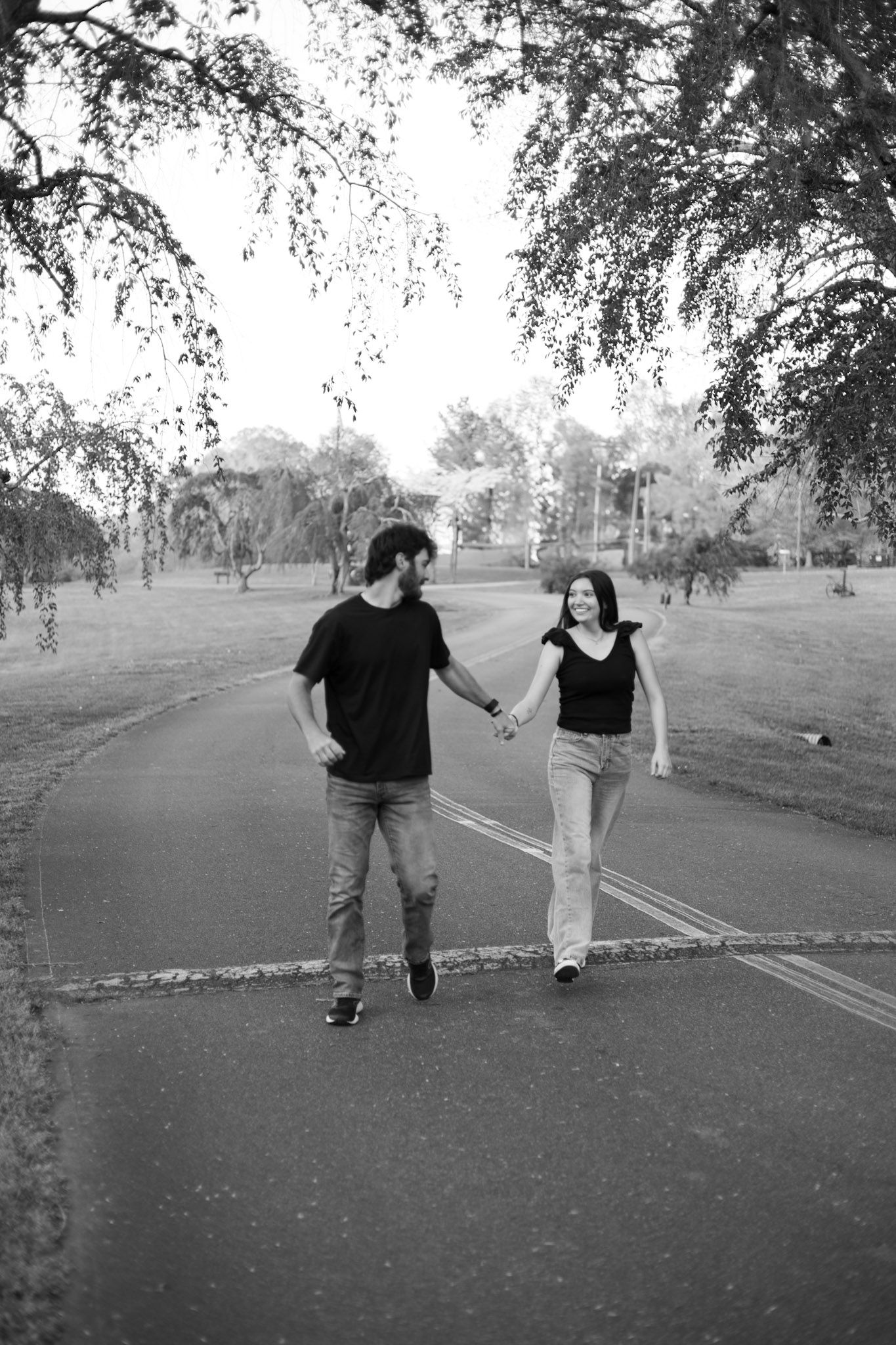 Two people hold hands while walking together on a paved path under drooping tree branches in black and white.