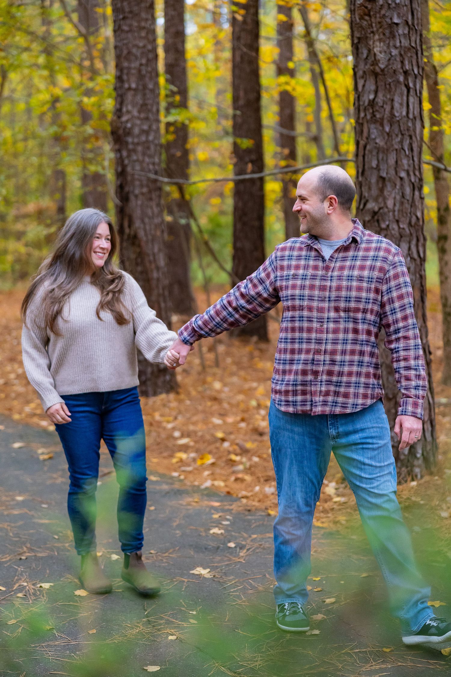 A couple walks together through a fall forest setting with golden leaves and pine trees in the background.