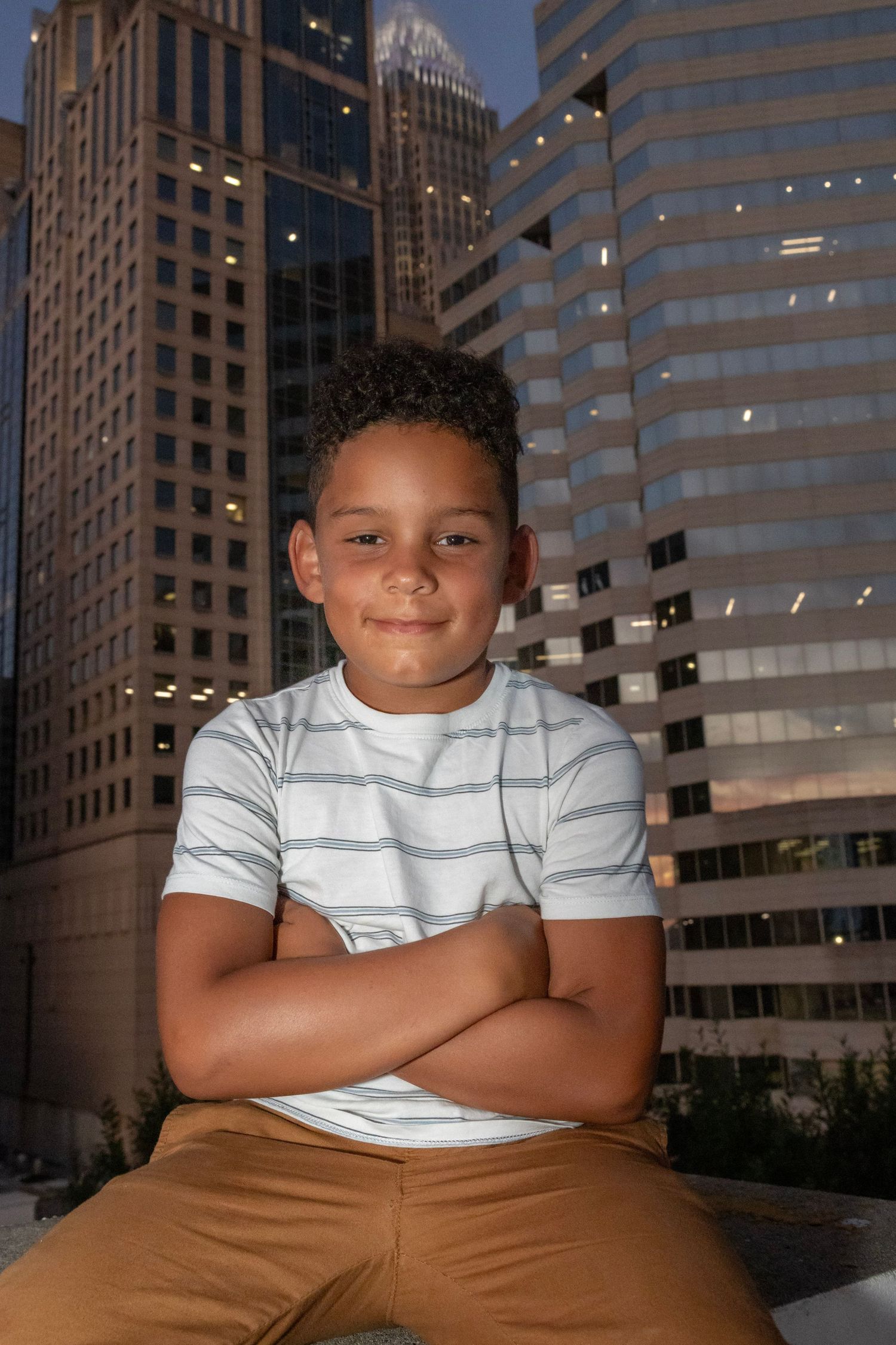 A young person in a striped shirt poses confidently against an urban backdrop of modern office buildings at dusk.