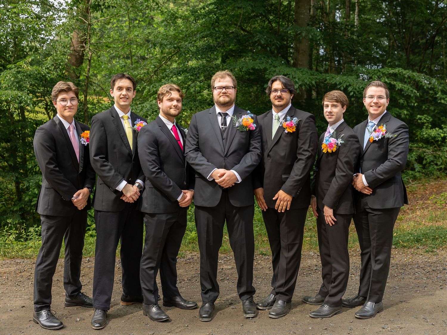 Seven groomsmen in black suits with colorful boutonnieres pose outdoors at a wedding.